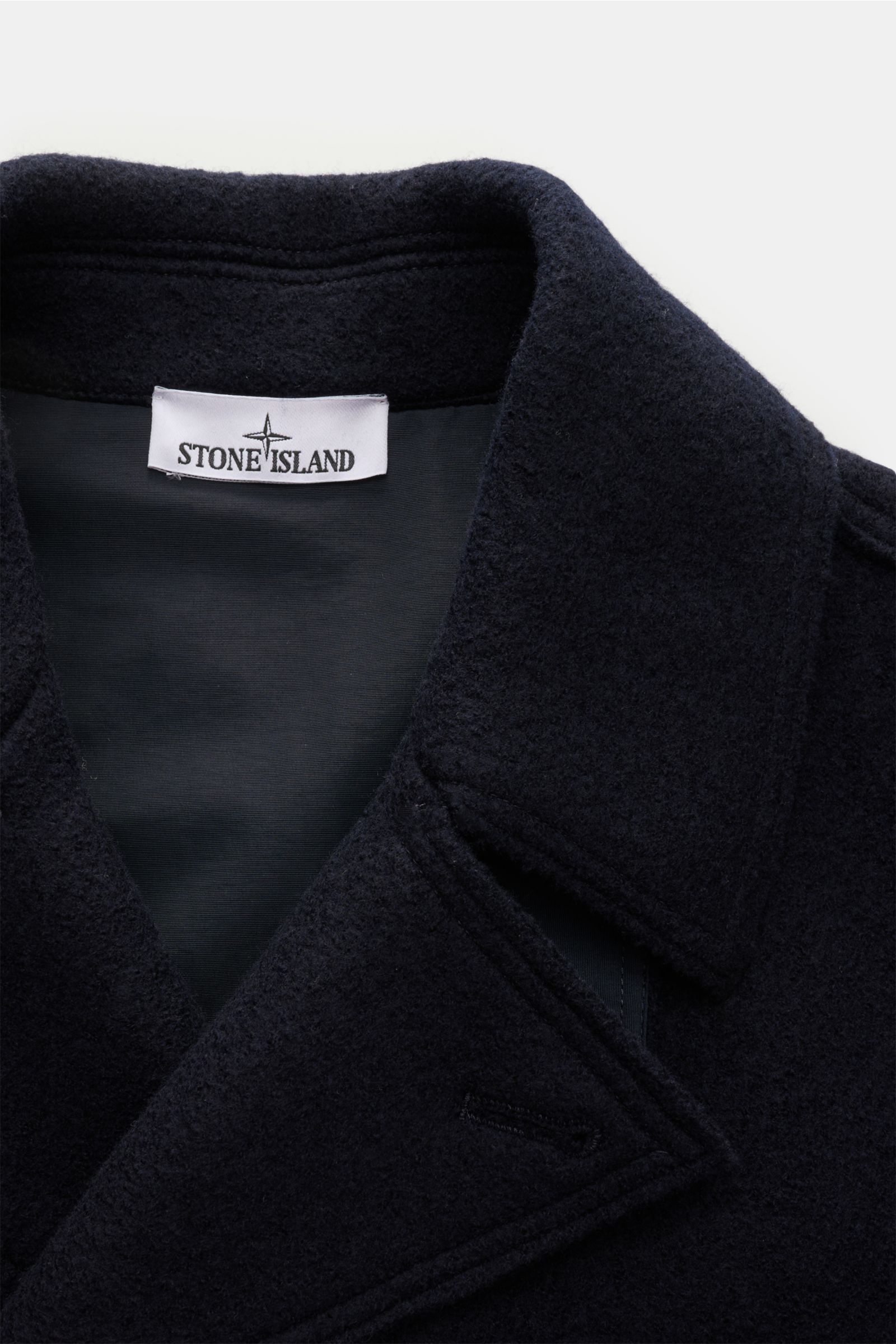Nahaufnahme der Stone Island Cabanjacke navy aus reiner, gefilzter Wolle mit weichem Griff, garment dyed, variabler Kragen mit Knopfriegel, zweireihiger Drei-Knopf-Leiste, schrägen Leistentaschen und typischem Kompass-Badge am Ärmel, von oben fotografiert