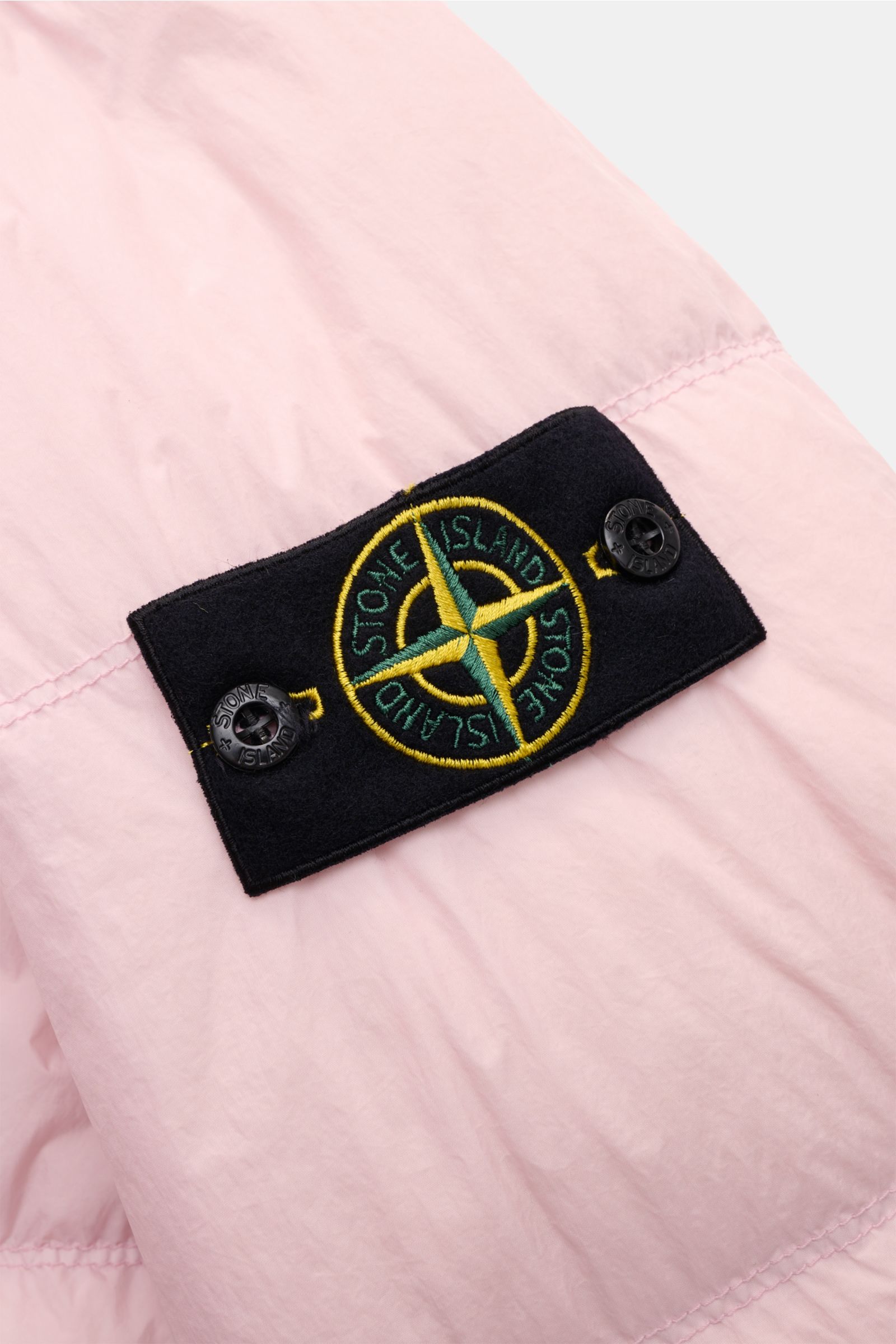 Close-up view of the Stone Island Daunenjacke 'Garment Dyed Crinkle Reps NY' rosé showing the black compass badge on the coated, garment-dyed pink microfiber fabric.  

Description: Bei der Daunenjacke 'Garment Dyed Crinkle Reps NY' von STONE ISLAND tr