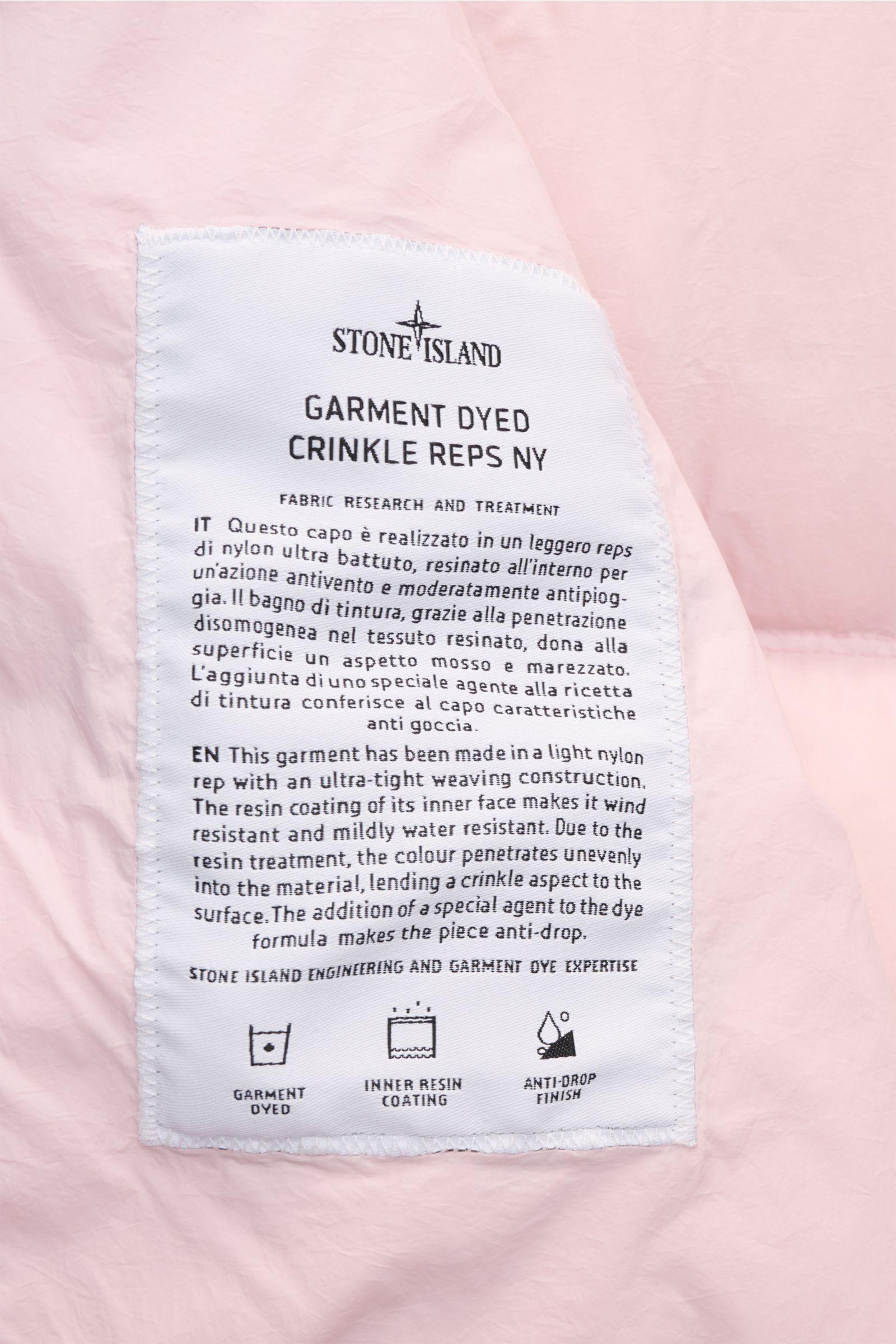 Close-up of the white fabric label sewn inside the Stone Island Daunenjacke 'Garment Dyed Crinkle Reps NY' rosé, photographed from above, showing detailed fabric research and treatment info.

Description: Bei der Daunenjacke 'Garment Dyed Crinkle Reps 