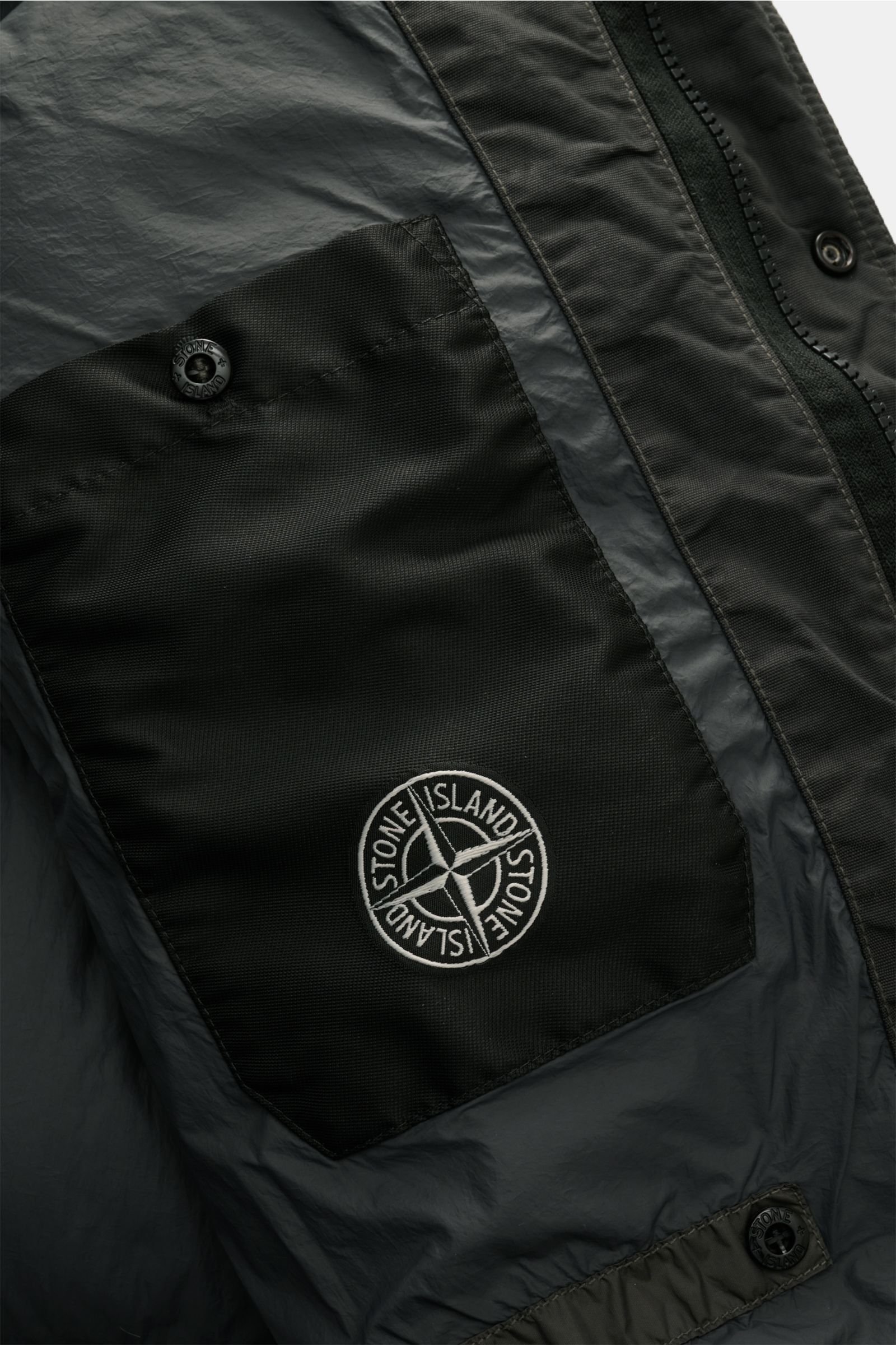 Close-up inside view of Stone Island Daunenjacke 'Garment Dyed Crinkle Reps NY' dunkelgrau showing black inner pocket with Stone Island compass badge and button details.

Description: Bei der Daunenjacke 'Garment Dyed Crinkle Reps NY' von STONE ISLAND 