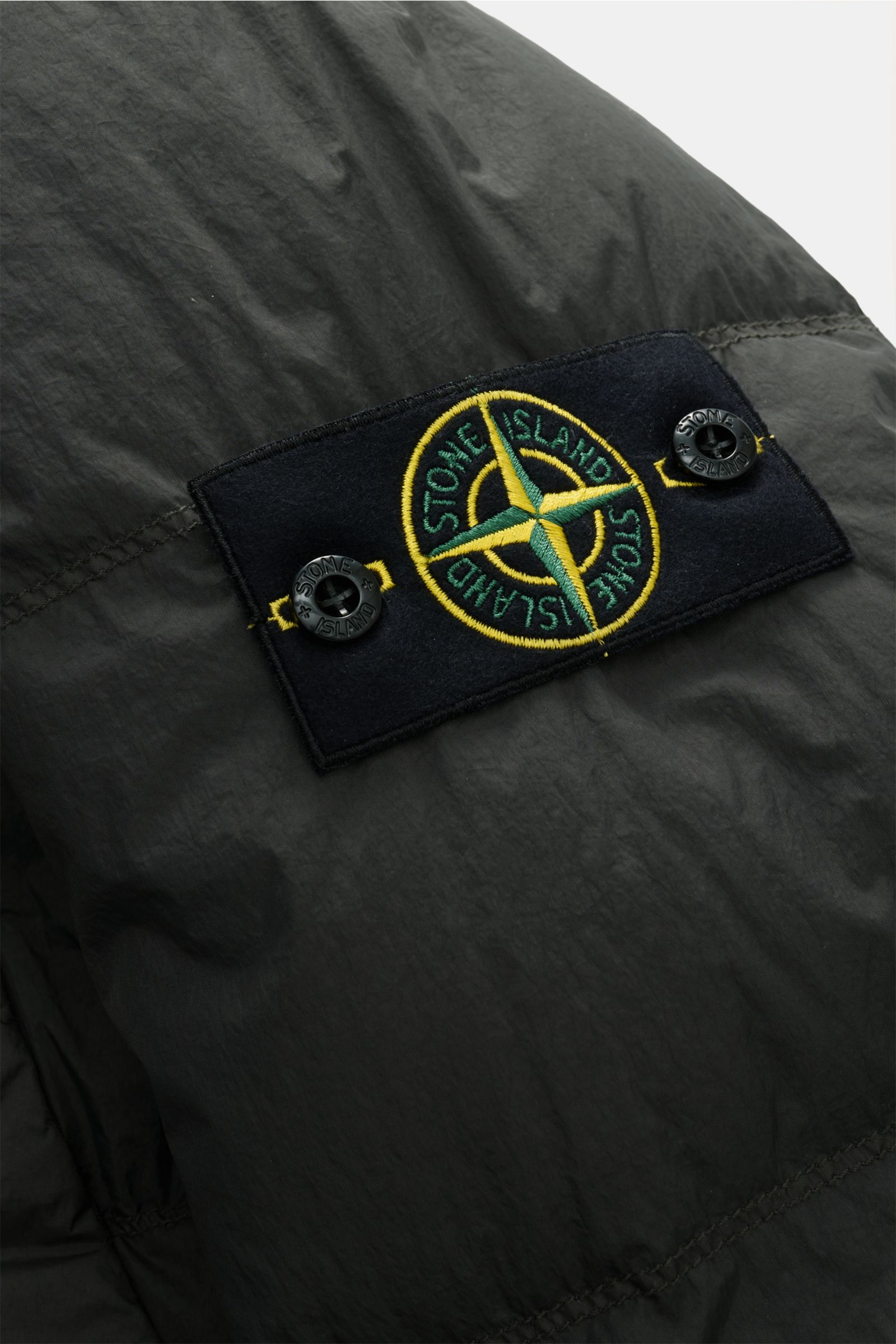 Close-up of the Stone Island Daunenjacke 'Garment Dyed Crinkle Reps NY' dunkelgrau showing the black patch with yellow and green compass logo, photographed from a side angle.

Bei der Daunenjacke 'Garment Dyed Crinkle Reps NY' von STONE ISLAND trifft b