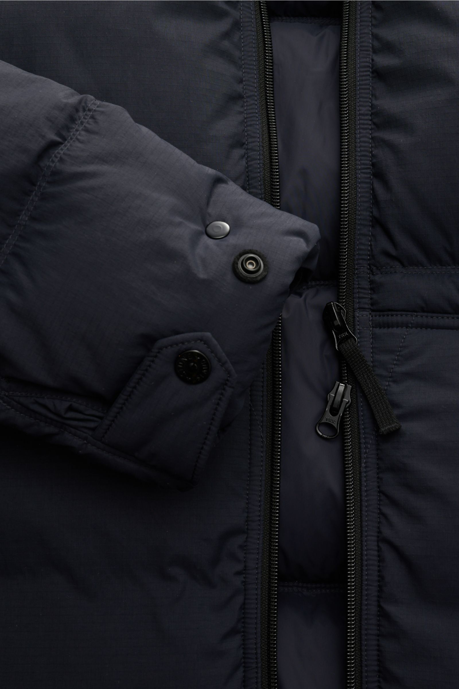 Nahaufnahme der Stone Island Daunenjacke 'Matt Ripstop Cordura' navy, zeigt wasserabweisendes Taslan-Gewebe, Reißverschluss und verstellbaren Ärmel mit Druckknopf, Slim Fit, Kapuze mit Tunnelzug, schräge Leistentaschen, Innentasche, wärmende Daunenfüllung