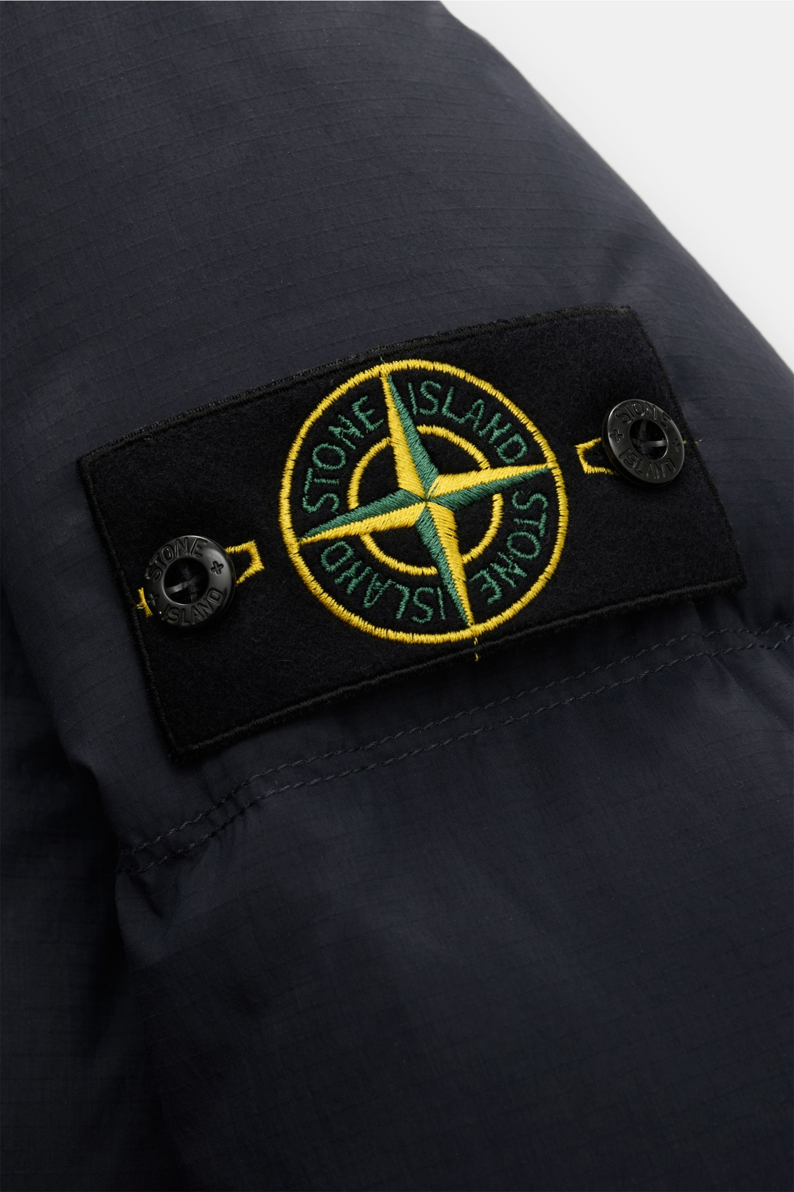 Nahaufnahme der Stone Island Daunenjacke 'Matt Ripstop Cordura' navy mit gesticktem Logo auf dem Ärmel, wasserabweisendem, mattem Taslan-Gewebe.