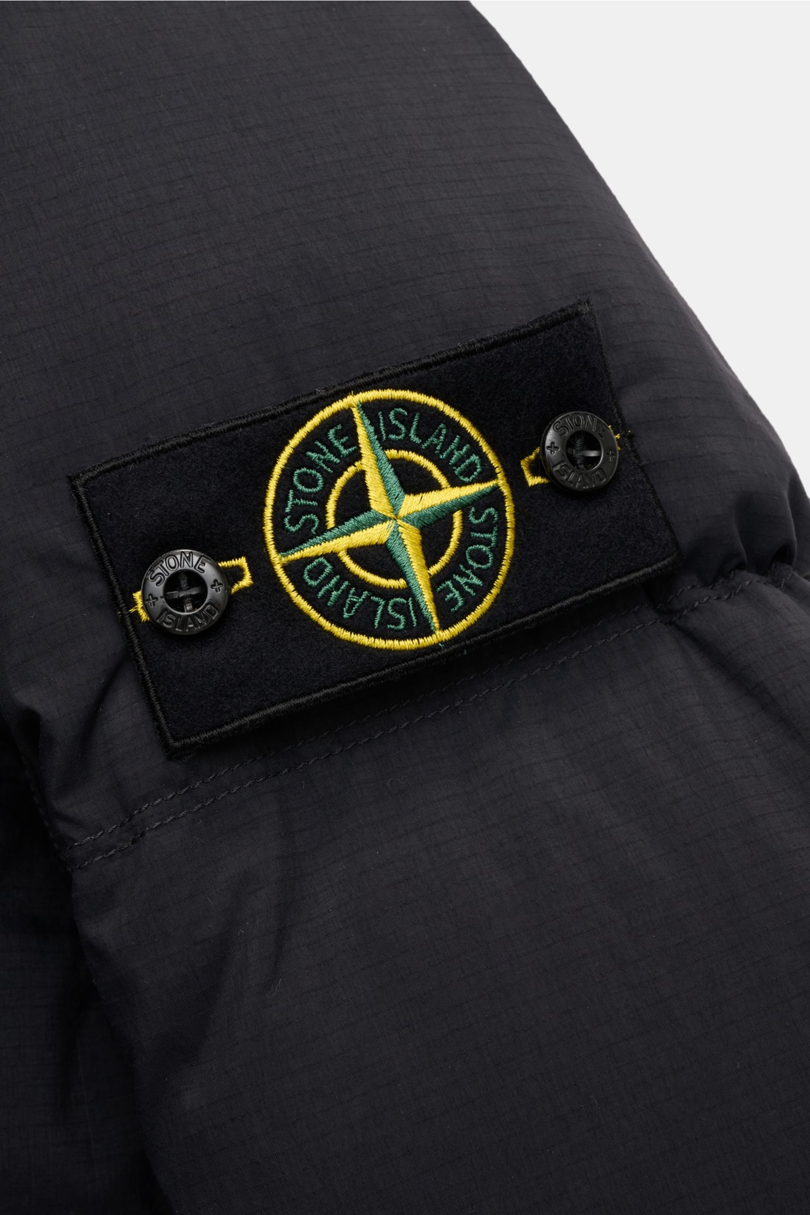 Close-up side view of the Stone Island Daunenjacke 'Matt Ripstop Cordura' navy showing the black patch with yellow-green compass logo on the sleeve. Legerer Look trifft Funktion: Die Daunenjacke von STONE ISLAND kombiniert sportiv-coole Ästhetik mit funkt