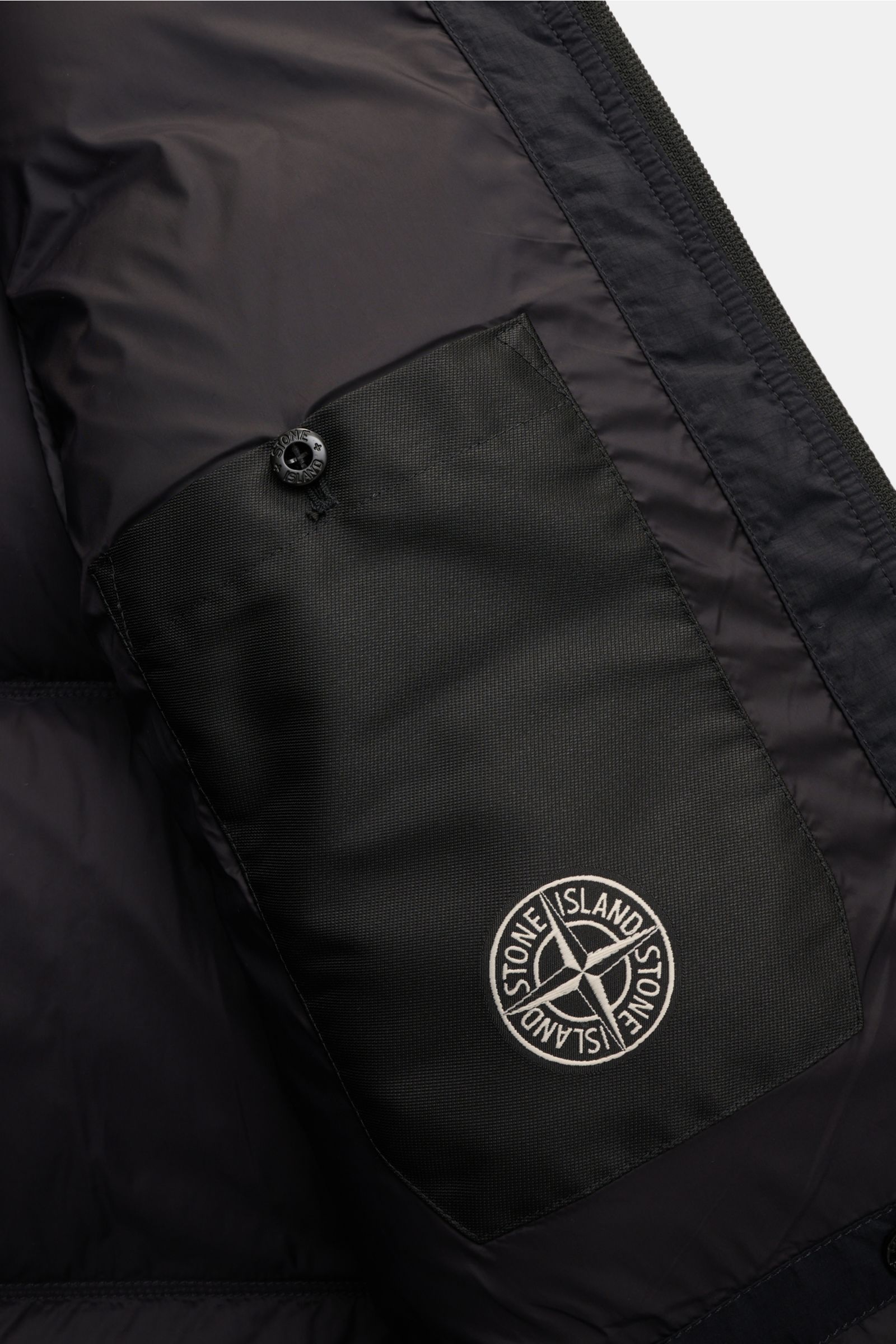Close-up inside view of the Stone Island Daunenjacke 'Matt Ripstop Cordura' navy showing the black inner pocket with button and embroidered compass badge. Legerer Look trifft Funktion: Die Daunenjacke von STONE ISLAND kombiniert sportiv-coole Ästhetik mit