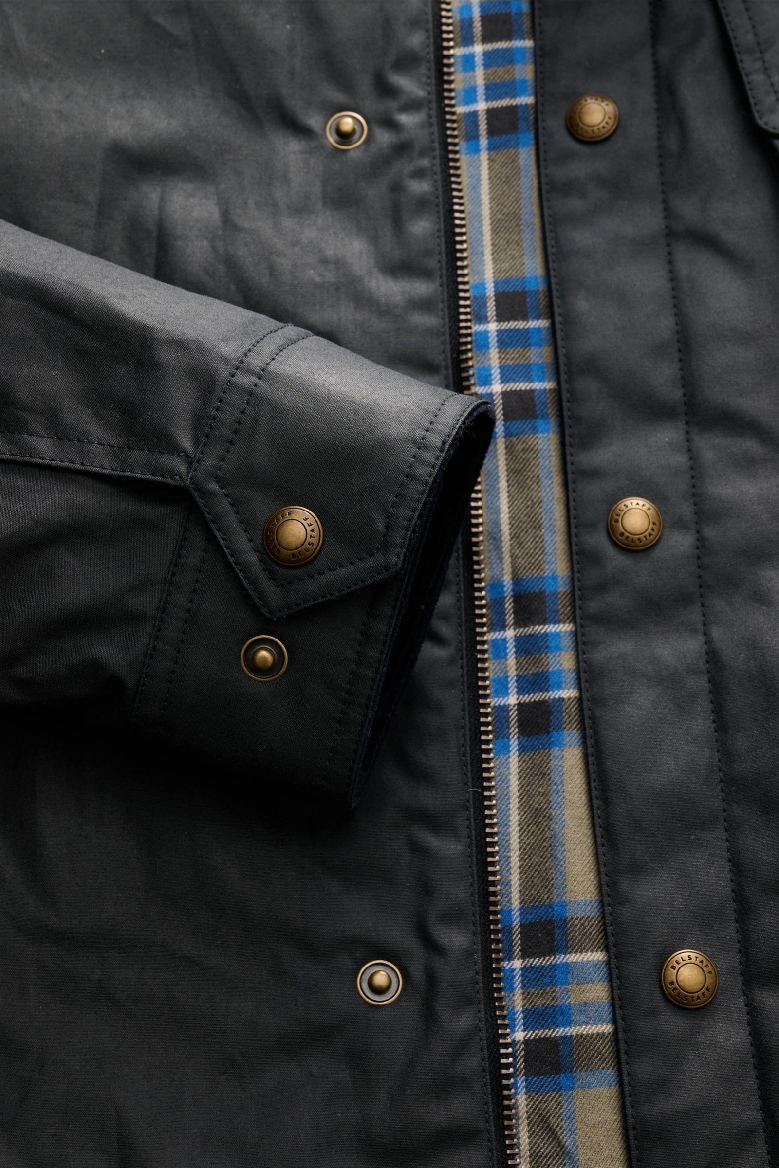 Nahaufnahme der Belstaff Jacke 'Ground' dark olive von oben fotografiert, zeigt gewachste Baumwolle, Slim Fit, Cordbesatz am Umlegekragen, robusten Reißverschluss, Windblende mit Druckknöpfen, Tartan-Innenfutter, große Brusttasche, Ärmel mit Druckknopf, B
