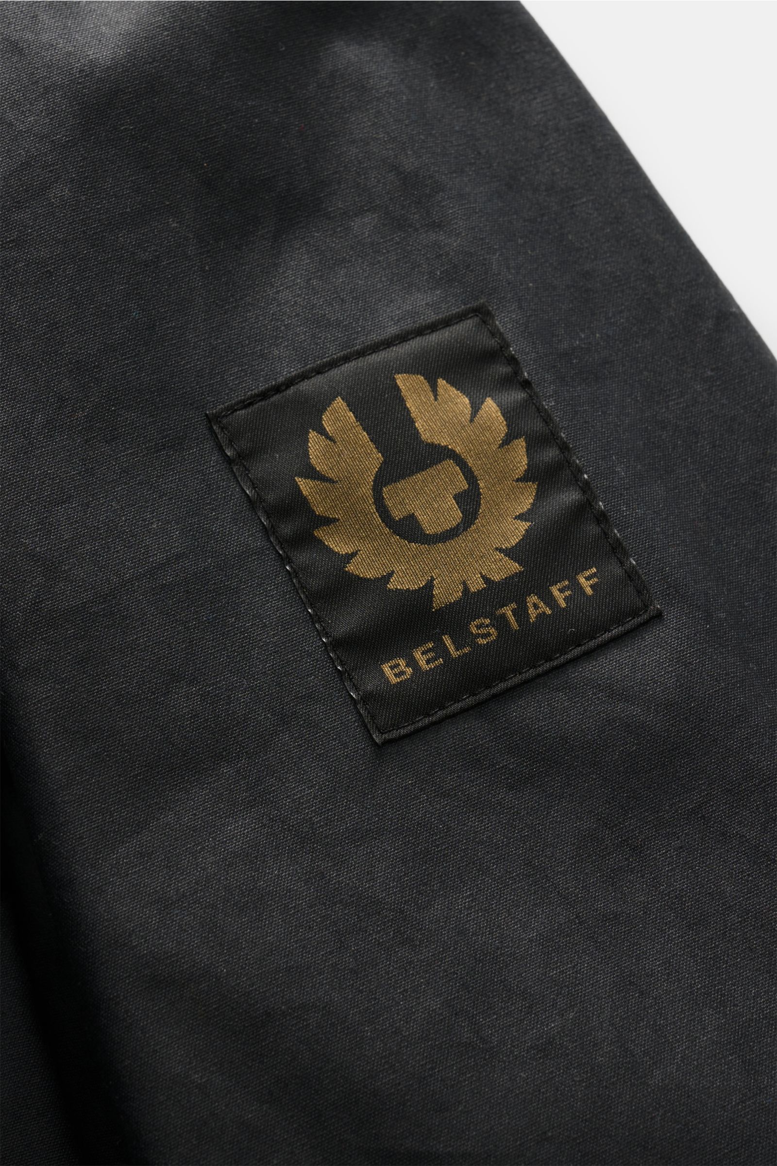 Nahaufnahme der Belstaff Jacke 'Ground' dark olive mit BELSTAFF-Weblabel am Oberarm aus gewachster Baumwolle, Detail im kastigen Schnitt.