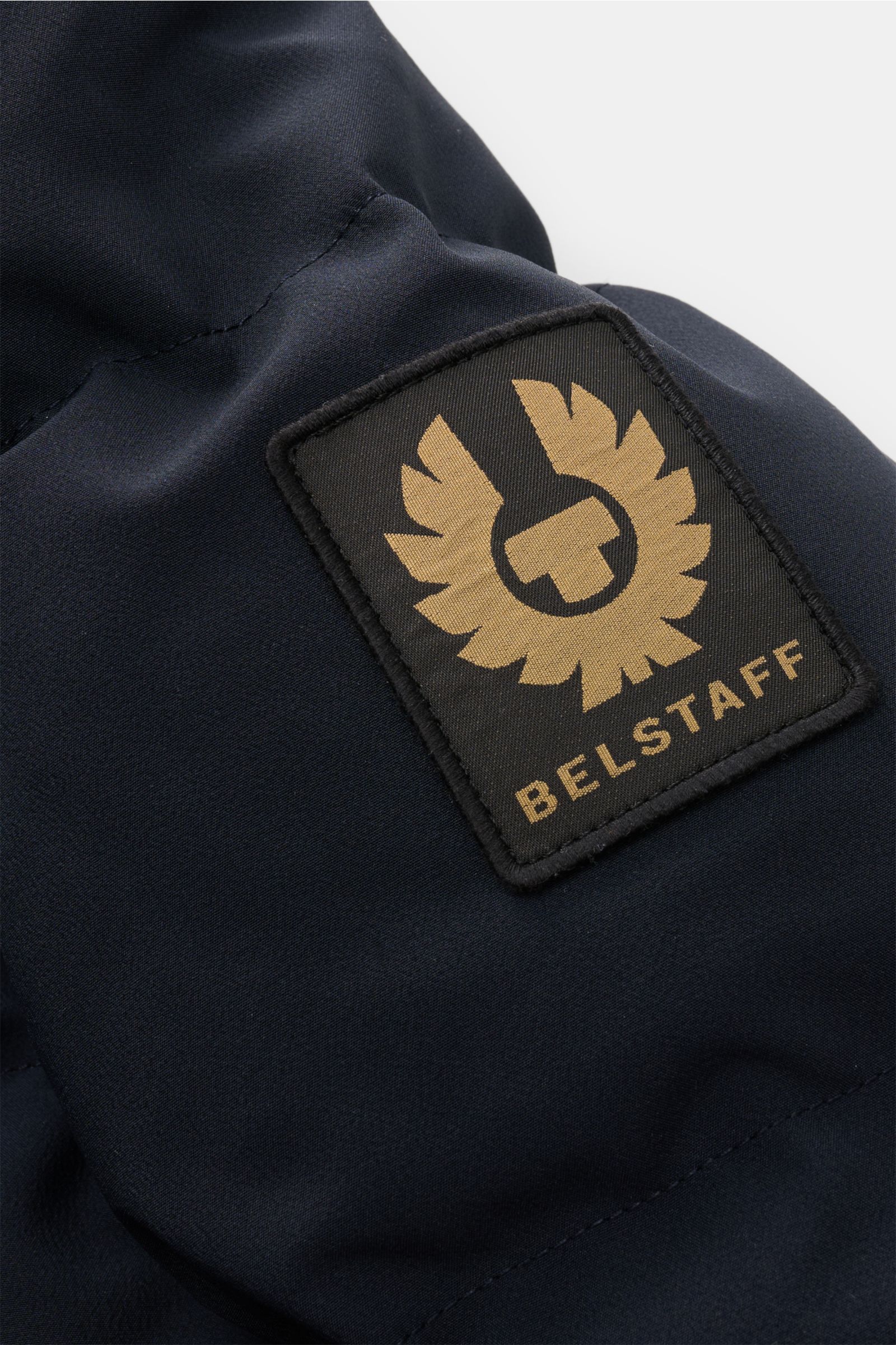 Nahaufnahme der Belstaff Daunenjacke 'Pendulum' navy von schräg oben, zeigt das schwarze BELSTAFF-Label auf hochwertiger, wind- und wasserabweisender Microfaser mit wärmender Daunenfüllung, abnehmbarer Kapuze und funktionalen Utility-Taschen.