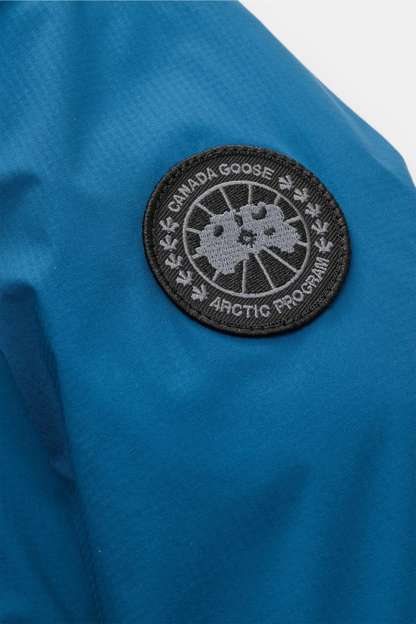 Blue ripstop fabric close-up with black and grey CANADA GOOSE Arctic Program patch, showing detail from a front-side perspective of the Canada Goose Down jacket 'Lodge Hoody' blue. Maximum comfort in a lightweight design: A padded hood and a down lining m
