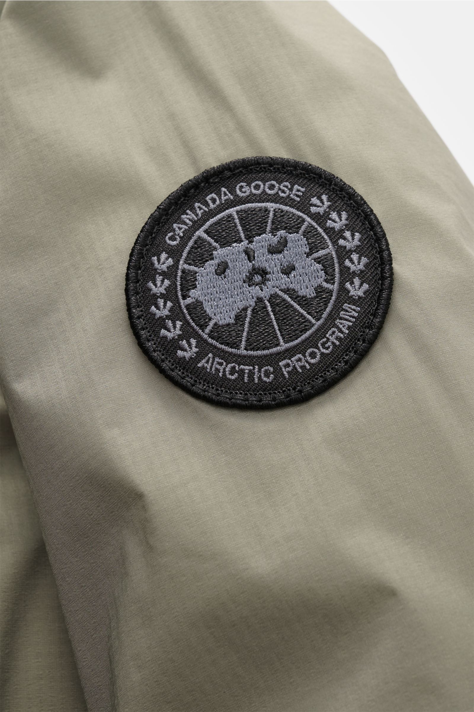 Canada Goose Daunenjacke 'Lodge Hoody' graugrün, Detailansicht des schwarzen CANADA GOOSE Weblabels auf leichtem Ripstop-Gewebe.