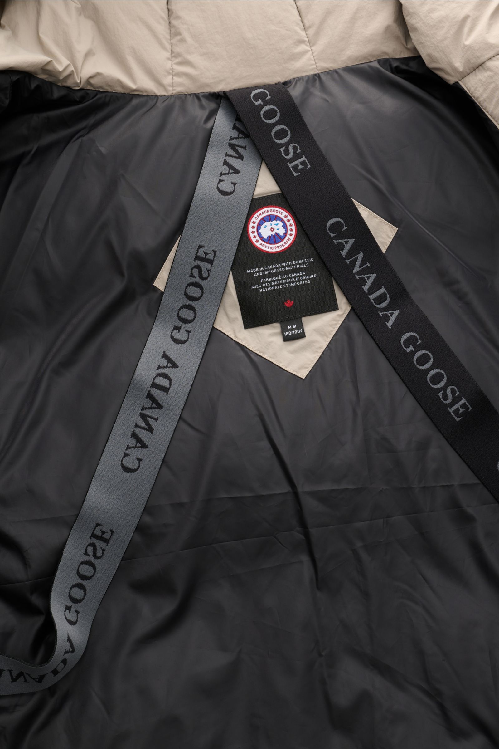 Innenansicht der Canada Goose Daunenjacke 'Crofton Puffer' sand mit glattem Futter, abnehmbaren Rucksackträgern und Labels, frontal fotografiert.