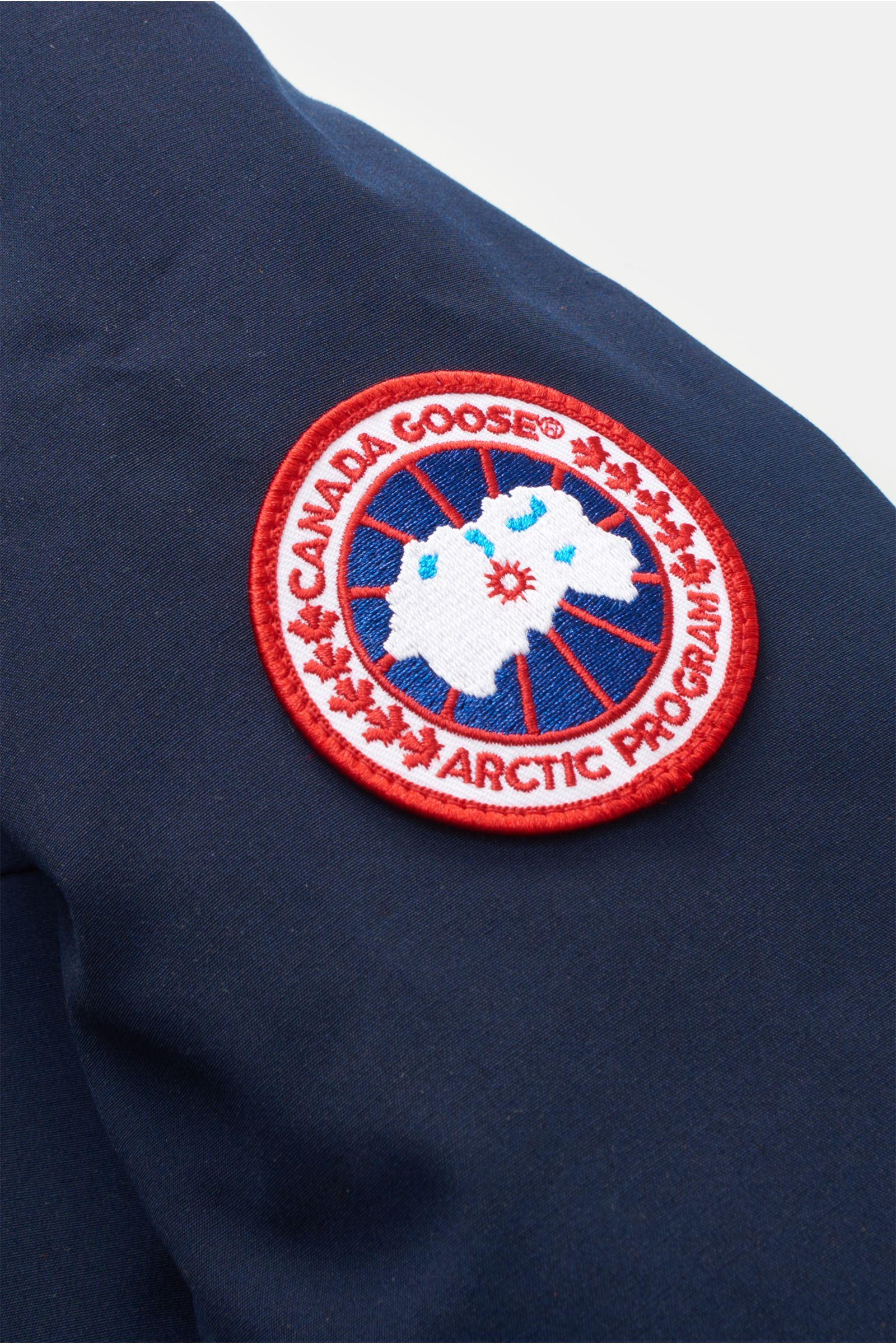 Nahaufnahme des roten Canada Goose Logos auf dem Ärmel des navy Canada Goose Daunenparka 'Langford' mit Arctic-Tech-Material.