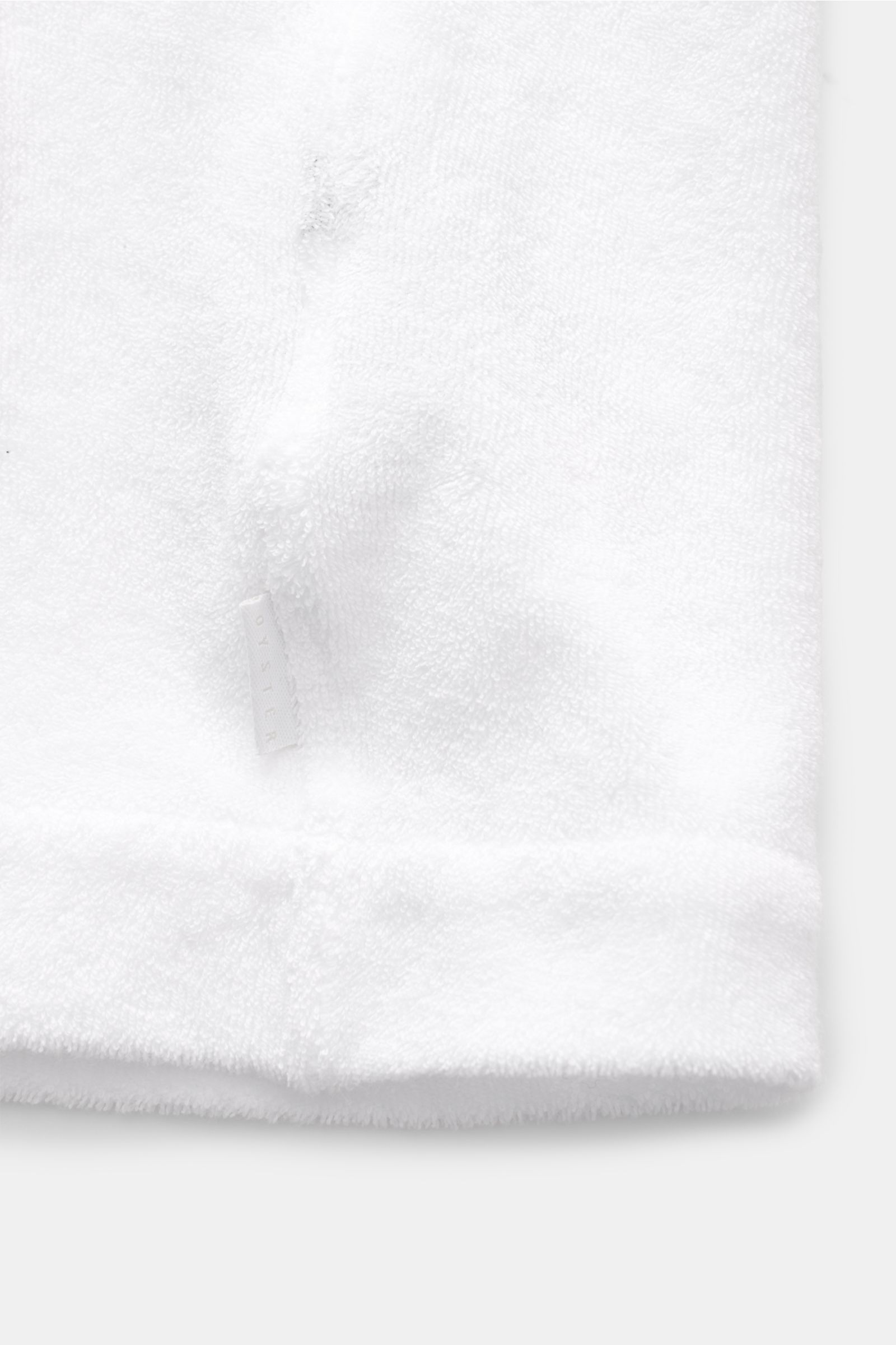 White terry fabric close-up showing texture and a small white label, photographed from above. The 04651/ A trip in a bag Frottee-Overshirt 'Terry Jacket' oliv features pure cotton terry, soft feel, slim fit, turn-down collar, five-button placket, patch fr