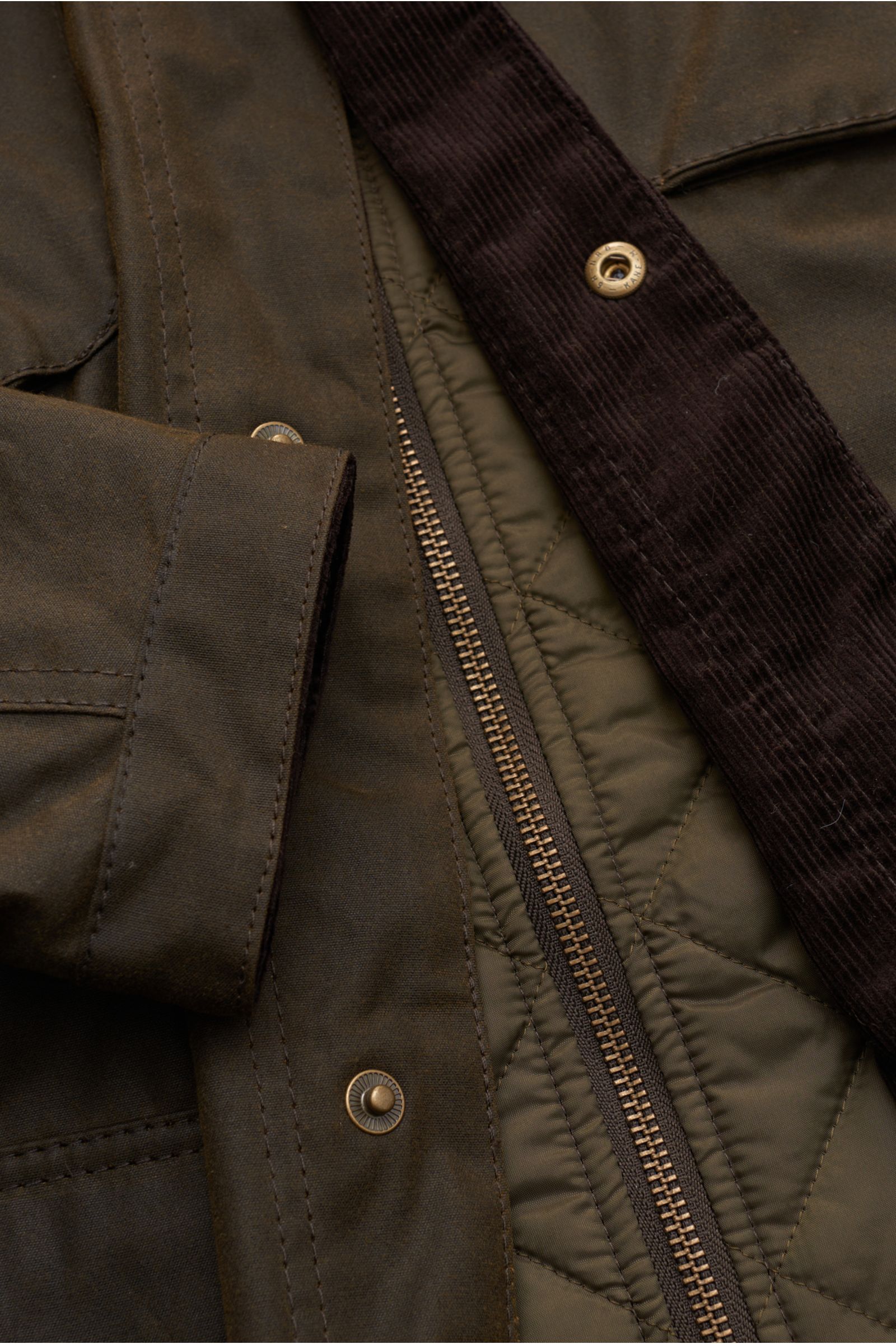 Close-up view of Barbour Waxed jacket 'Ogston' dark olive showing waxed cotton fabric, quilted double-front lining, zipper, snap buttons, and corduroy collar detail.