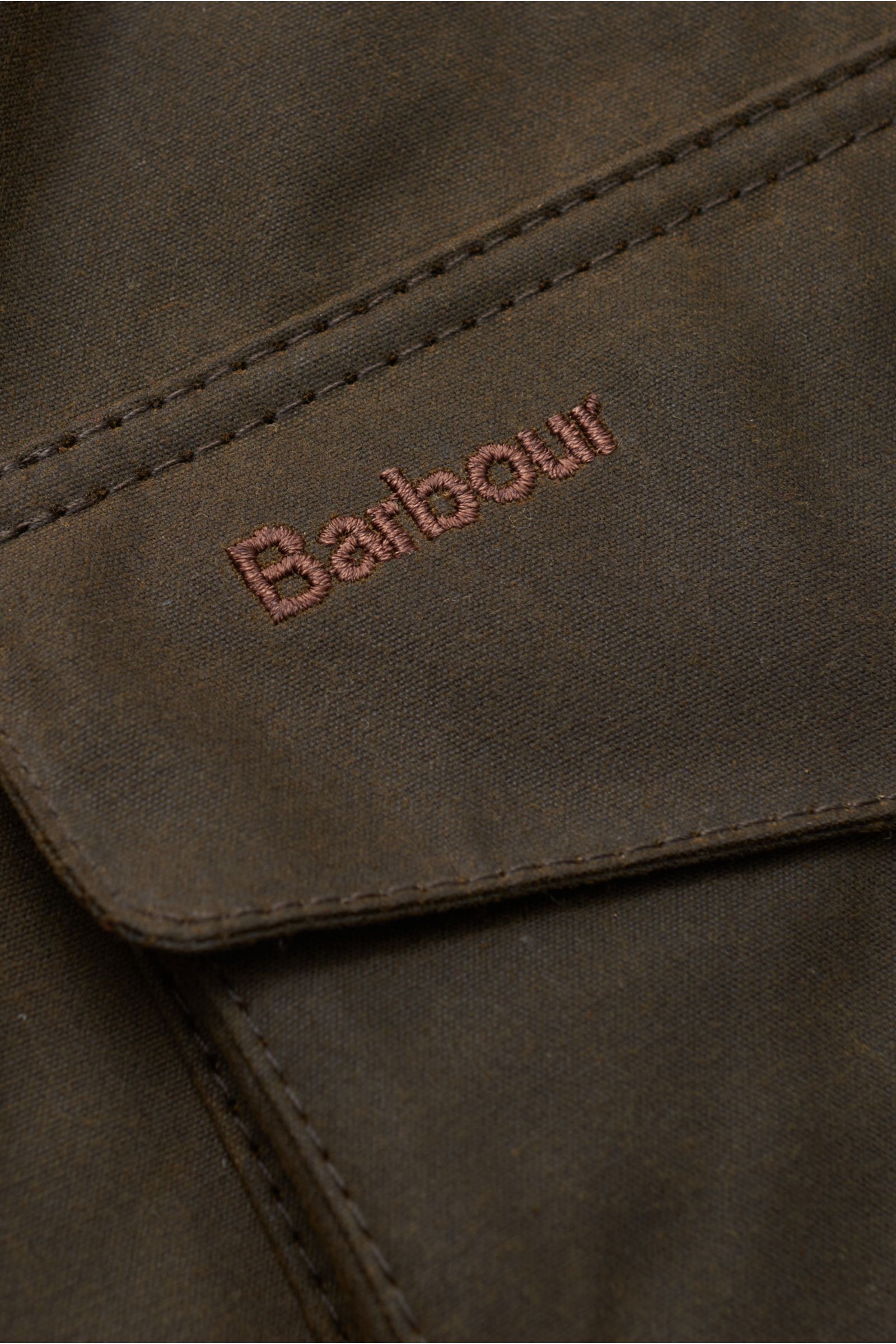 Close-up of Barbour Waxed jacket 'Ogston' dark olive showing the waxed cotton fabric and embroidered logo on a front pocket flap.

Elegant, stylish, masculine: The modern waxed jacket 'Ogston' by BARBOUR in water repellent, waxed cotton with double fro