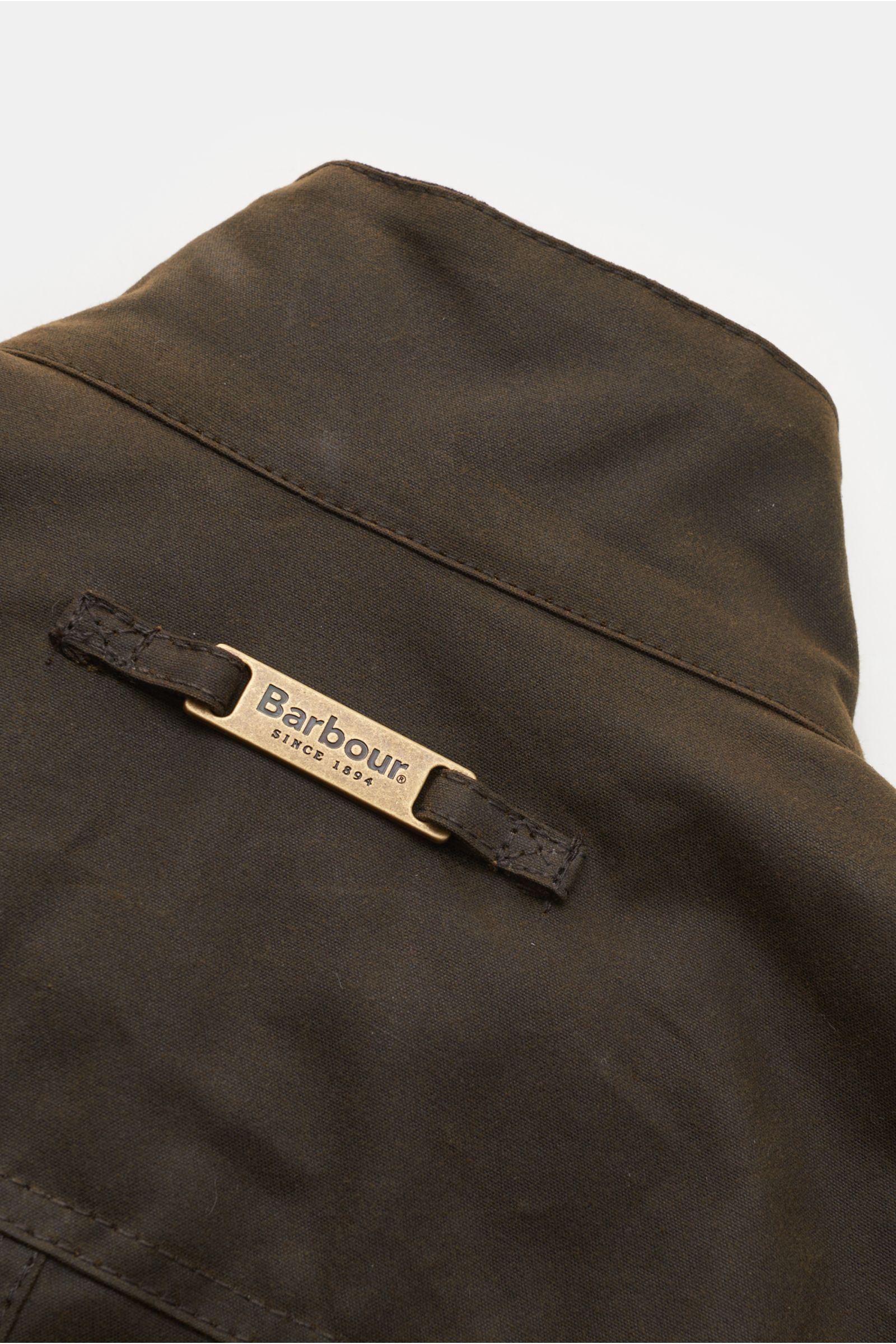 Close-up rear collar view of the Barbour Waxed jacket 'Ogston' dark olive, showing the signature Barbour metal logo detail on water repellent waxed cotton. Elegant, stylish, masculine: The modern waxed jacket 'Ogston' by BARBOUR in water repellent, waxed 