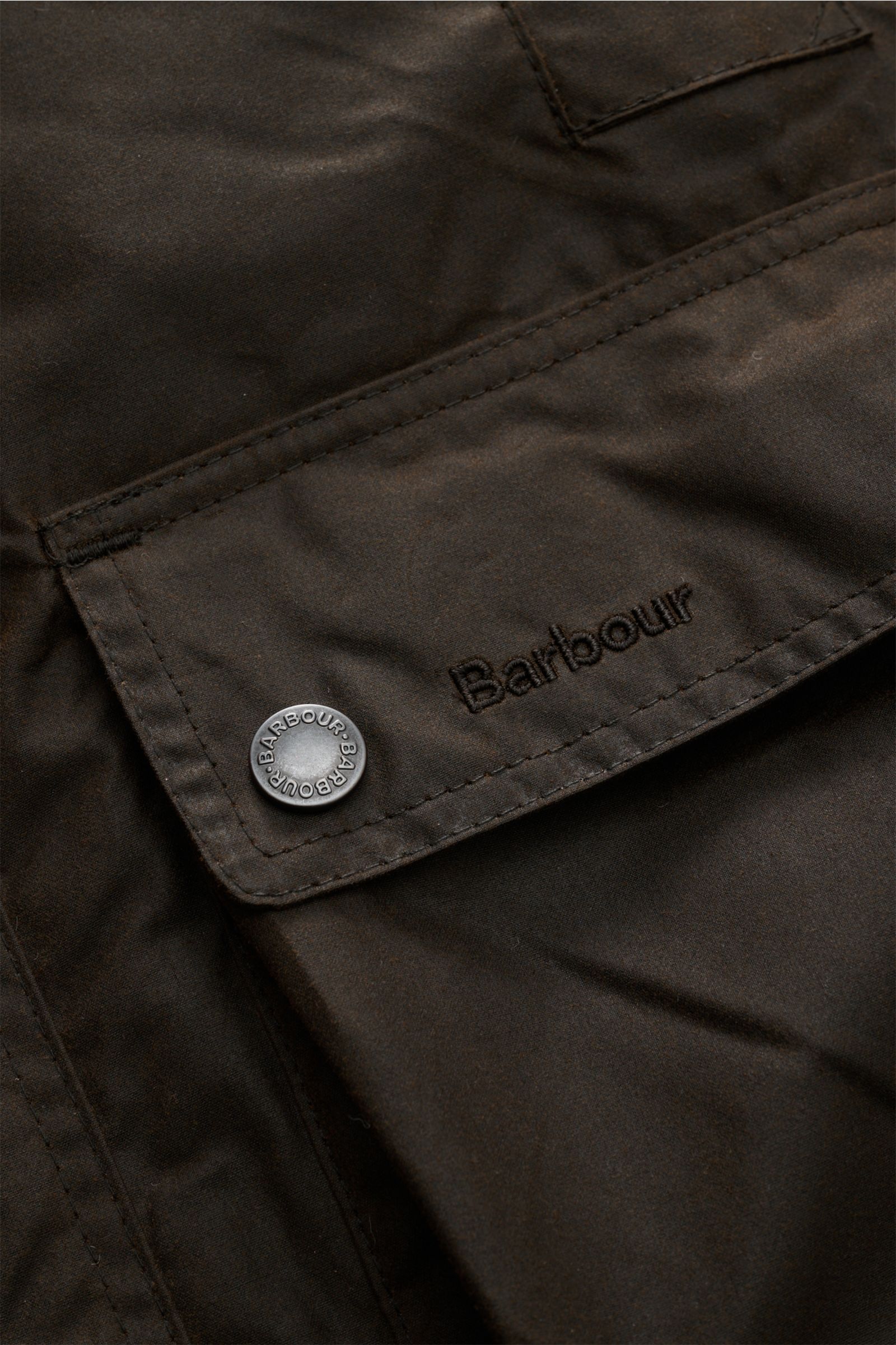 Nahaufnahme der Barbour Wachsjacke 'Modern Beaufort' dark olive mit gewachster Baumwolle, aufgesetzter Fronttasche und Logo-Detail.