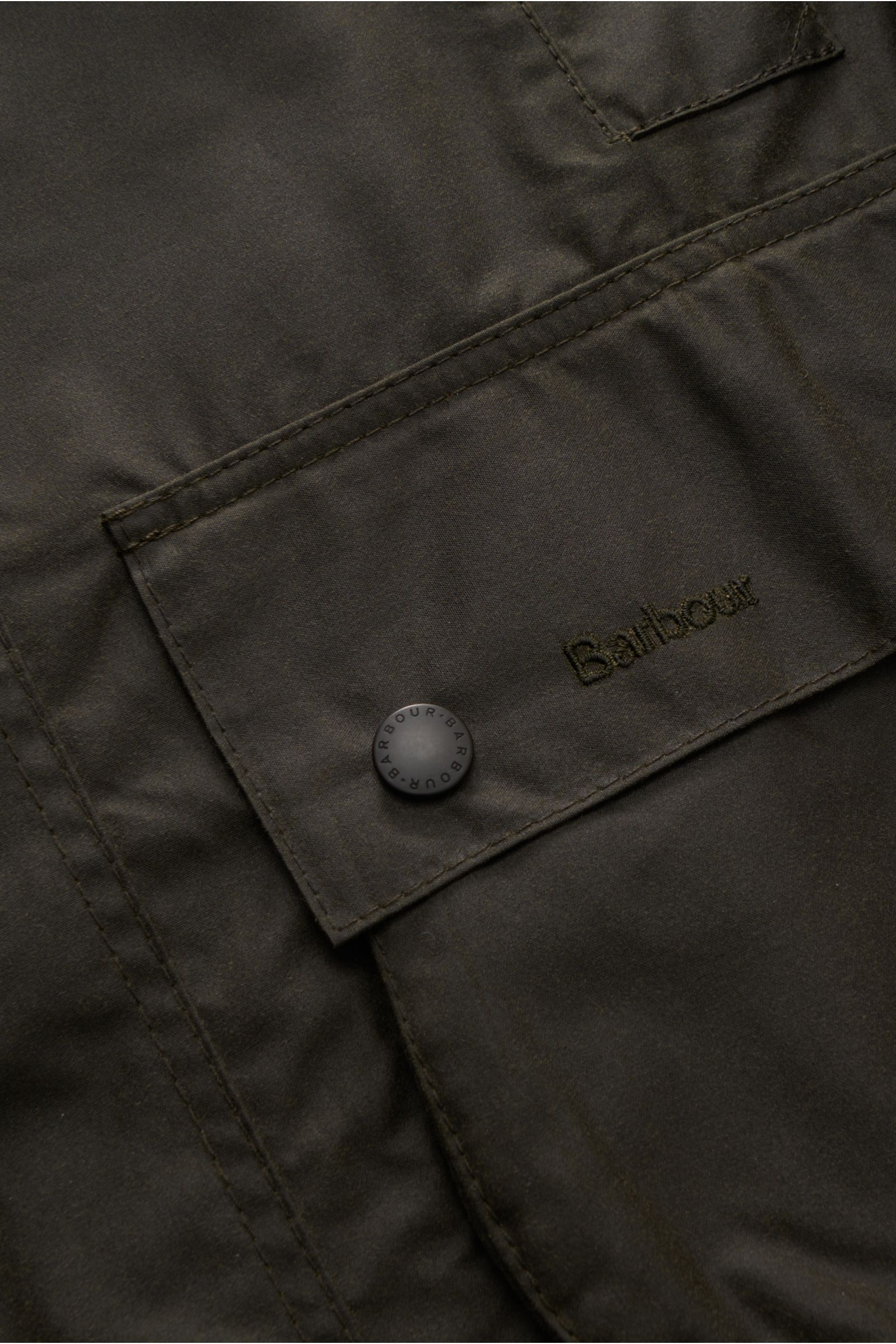 Close-up of the front pocket of Barbour Waxed jacket 'Beaufort' dark olive, showing waxed cotton fabric, snap button, and Barbour embroidery, photographed from a top-down perspective. A true classic from BARBOUR: The long waxed jacket 'Beaufort' made of w