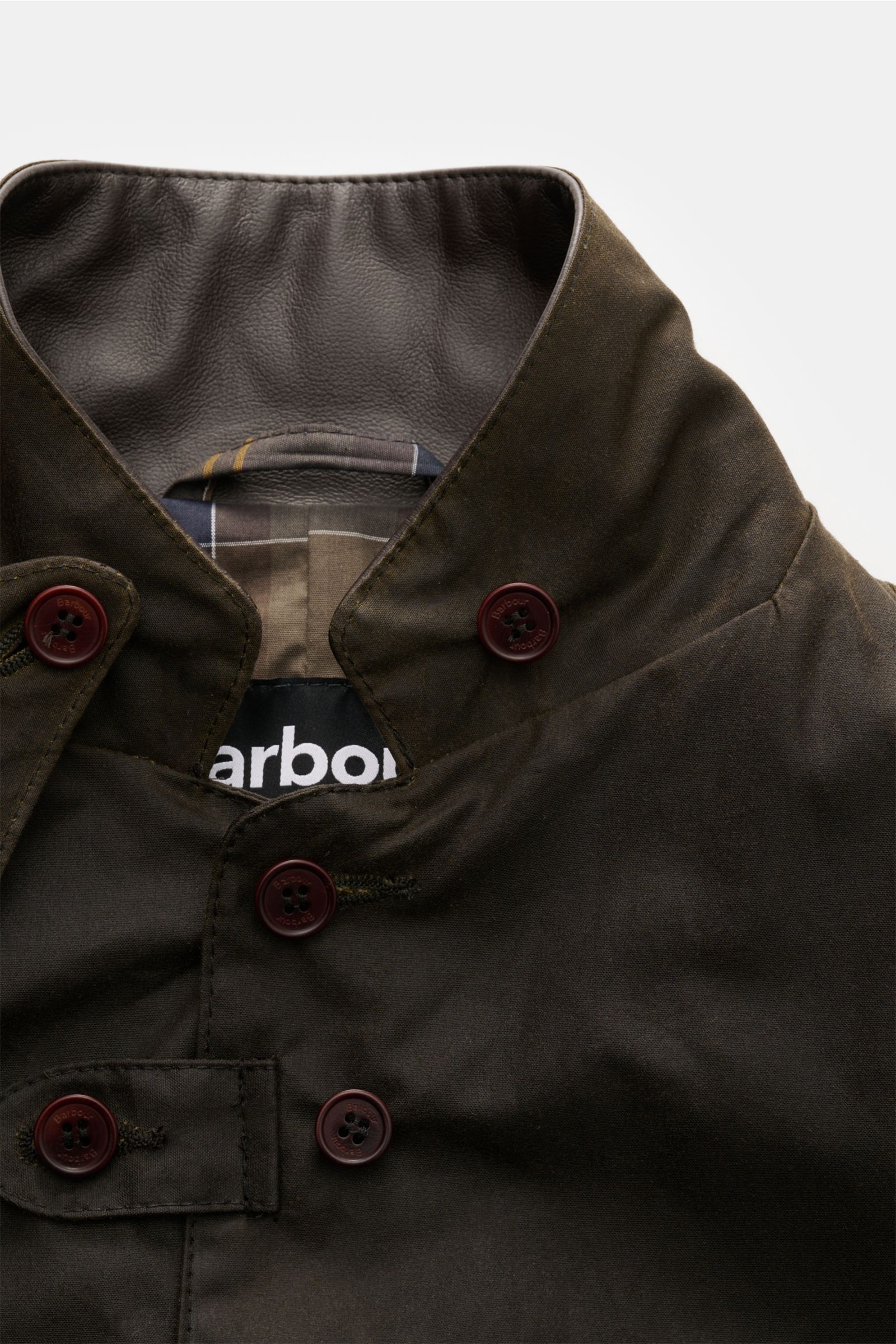 Barbour Wachsjacke 'Becan' dark olive in Nahaufnahme von oben, gewachste Baumwolle, Knopfleiste, Leder-Kragen, Tartan-Futter sichtbar.