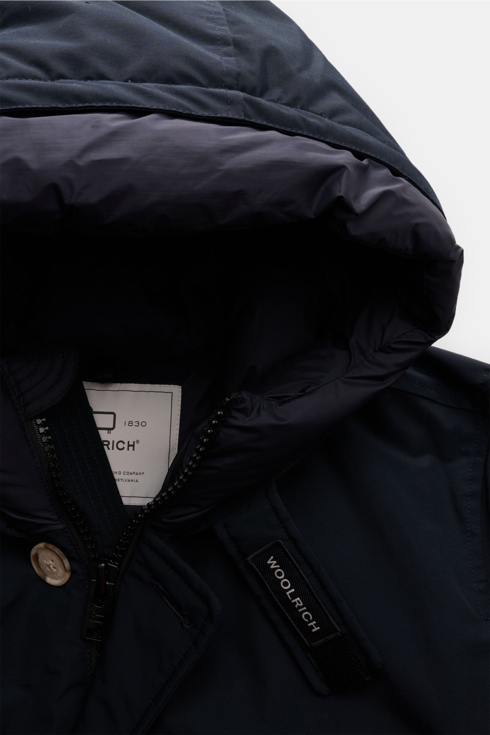 Close-up from above of the navy Woolrich Daunenparka 'Arctic Parka' showing the hood interior, zipper, button, and Woolrich label. Kälte? Kann kommen. Der 'Arctic Parka' von WOOLRICH liefert sportiven Style und verlässliche Wärme bis –20 °C – dank hochwer