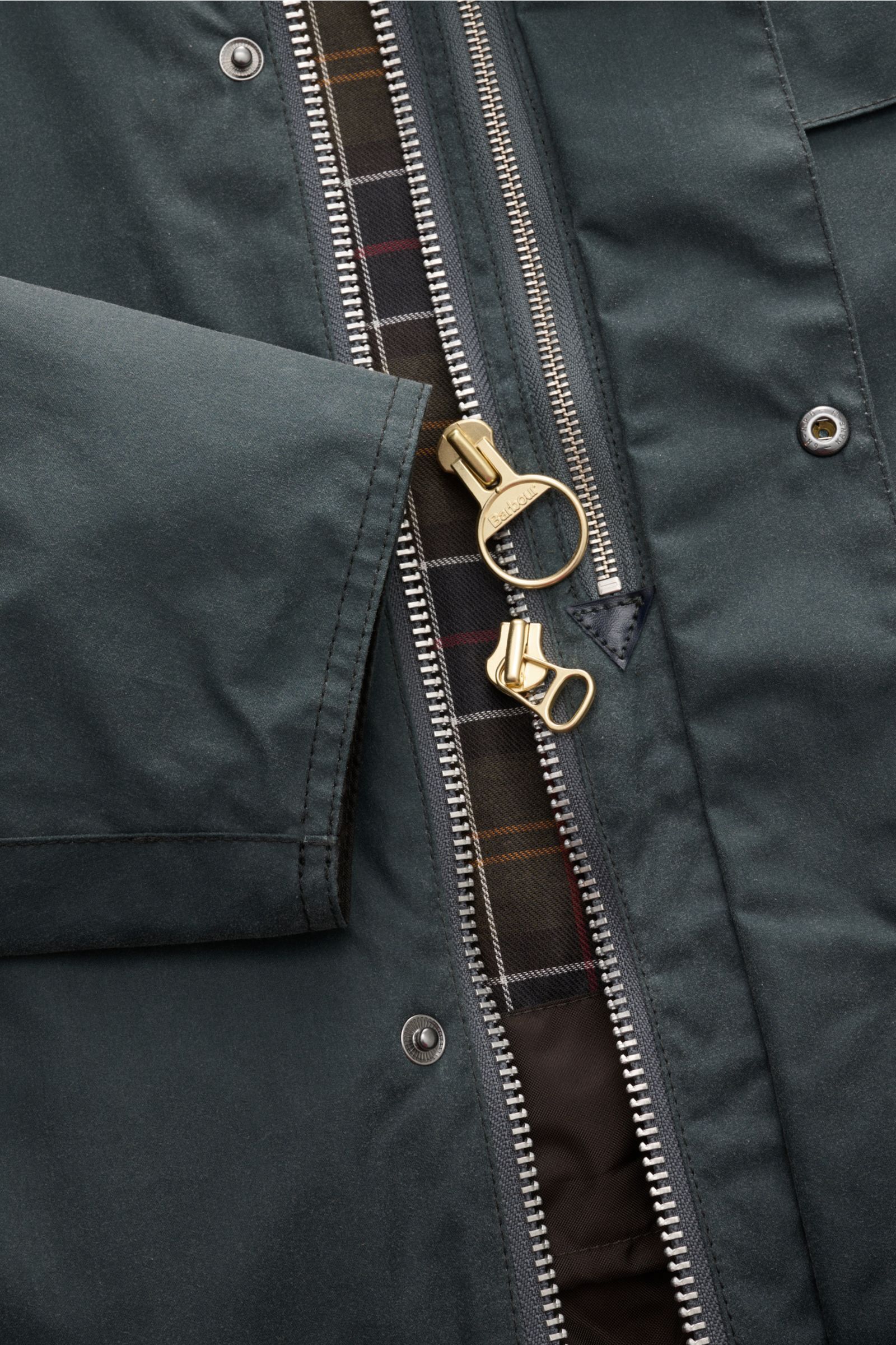 Nahaufnahme der Barbour Wachsjacke 'Transport' petrol von vorne, zeigt gewachste Baumwolle, doppelten Reißverschluss, Tartan-Innenfutter mit Vogel-Prints und Cord-Kragen.