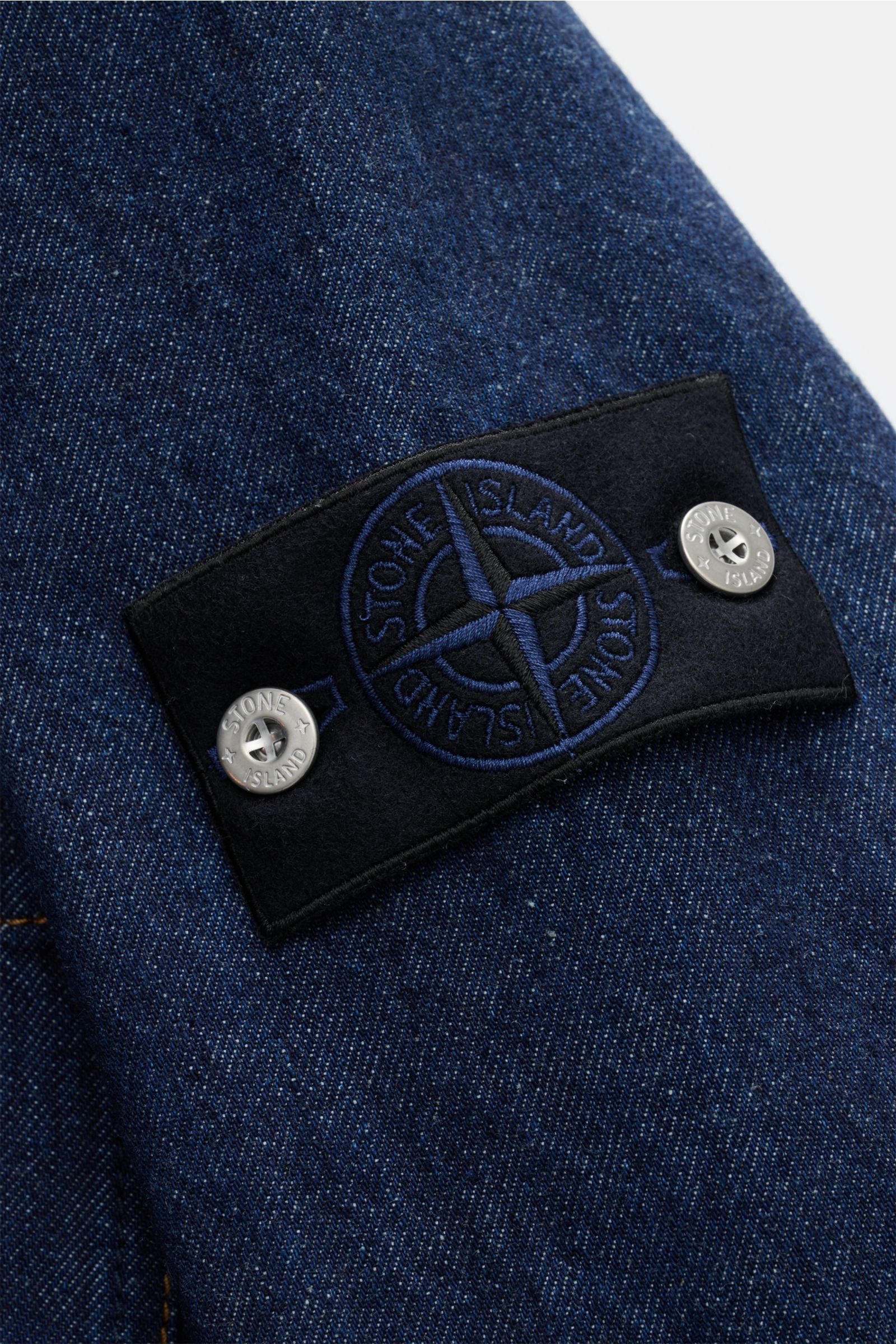 Stone Island Jeansjacke 'Indigo Denim-Rinsed' dunkelblau aus Baumwolle mit Kompass-Badge am Ärmel, Detailfoto schräg von oben.