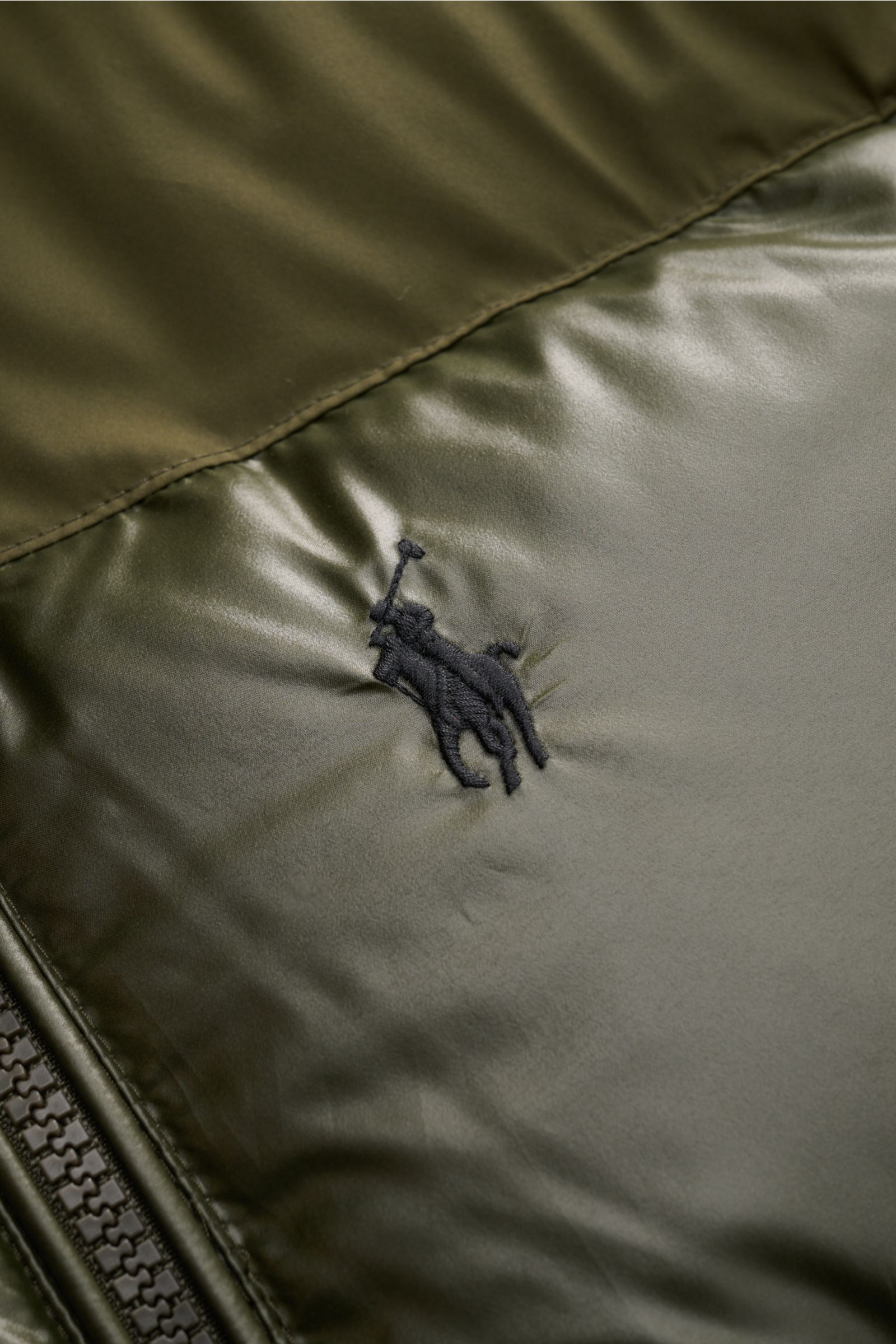 Close-up front view of Polo Ralph Lauren Daunenjacke oliv showing shiny olive fabric with black embroidered logo, smooth texture, and zipper details. Setzen Sie ein Statement - auch im Winter: Diese Daunenjacke von POLO RALPH LAUREN wärmt dank ihrer Da