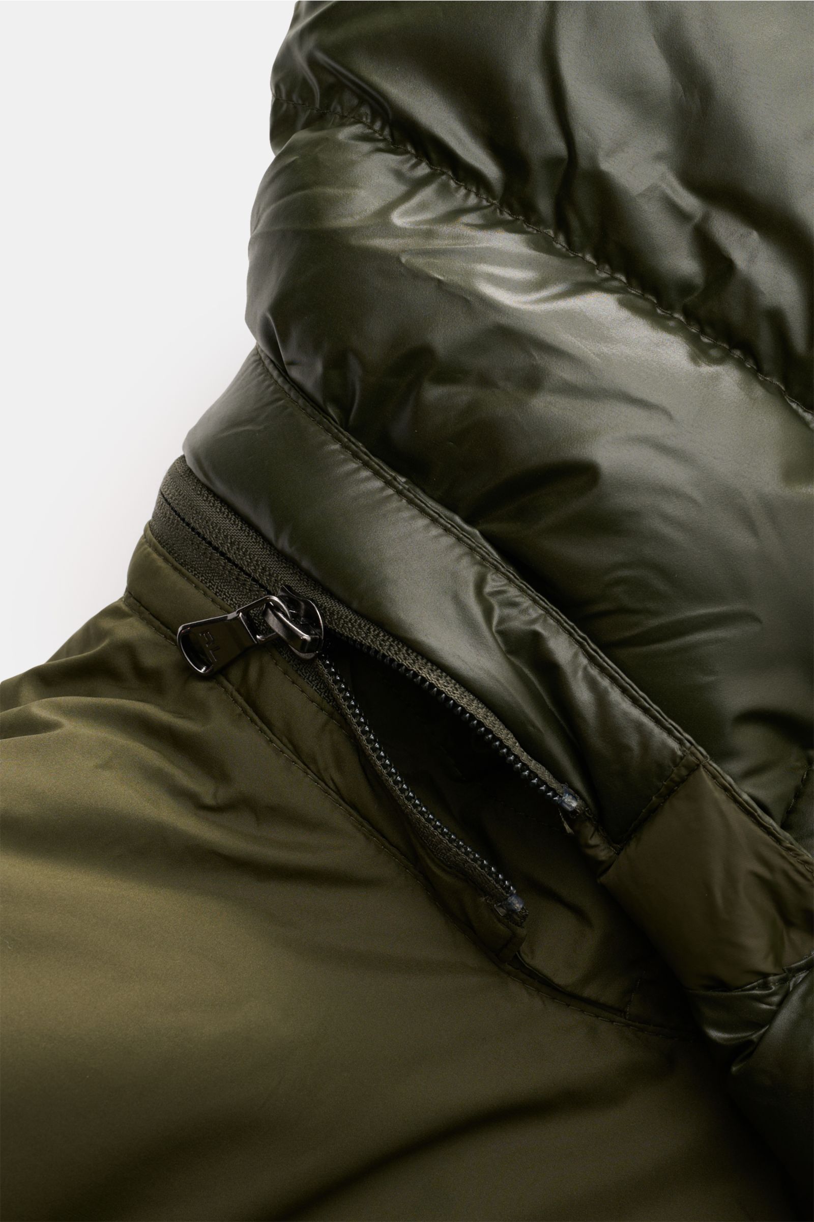 Close-up image of the zipper and shiny fabric detail of the Polo Ralph Lauren Daunenjacke oliv, photographed from a side perspective. Setzen Sie ein Statement - auch im Winter: Diese Daunenjacke von POLO RALPH LAUREN wärmt dank ihrer Daunenfüllung gara