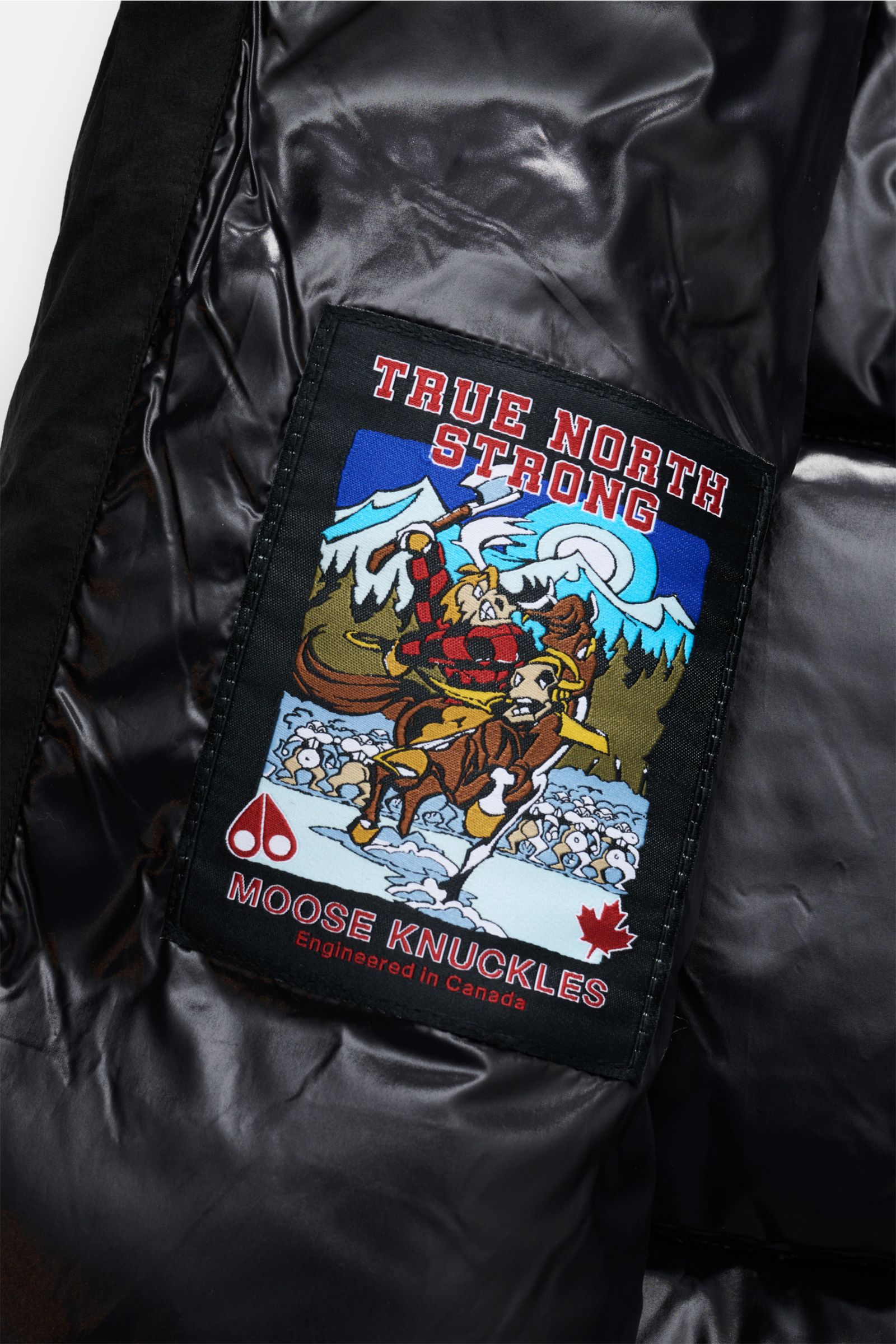 Close-up interior view of the Moose Knuckles Daunenjacke 'Everest 3Q' schwarz showing a colorful patch with 'True North Strong' text, moose and snowy mountain design on shiny black lining.

Höchster Komfort bei jeder Temperatur: Die Daunenjacke von MOO