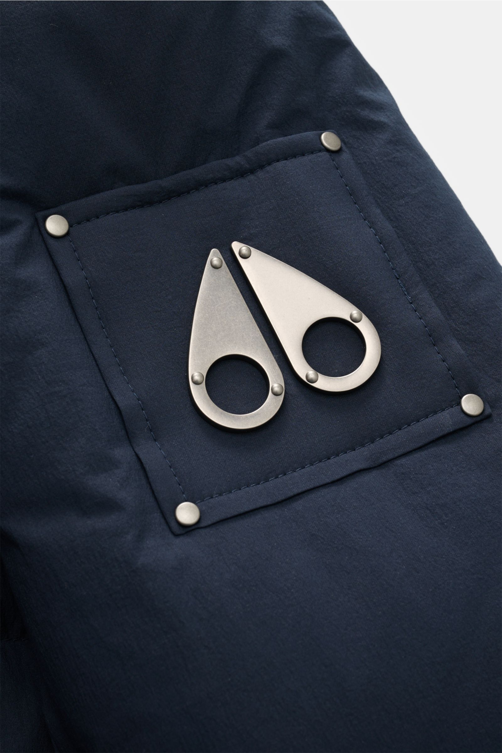 Nahaufnahme der Metall-Logoplakette an der Ärmeltasche der Moose Knuckles Daunenjacke 'Everest 3Q' navy, wind- und wasserabweisend, mit wärmender Daunenfüllung für Komfort bis -20 Grad.