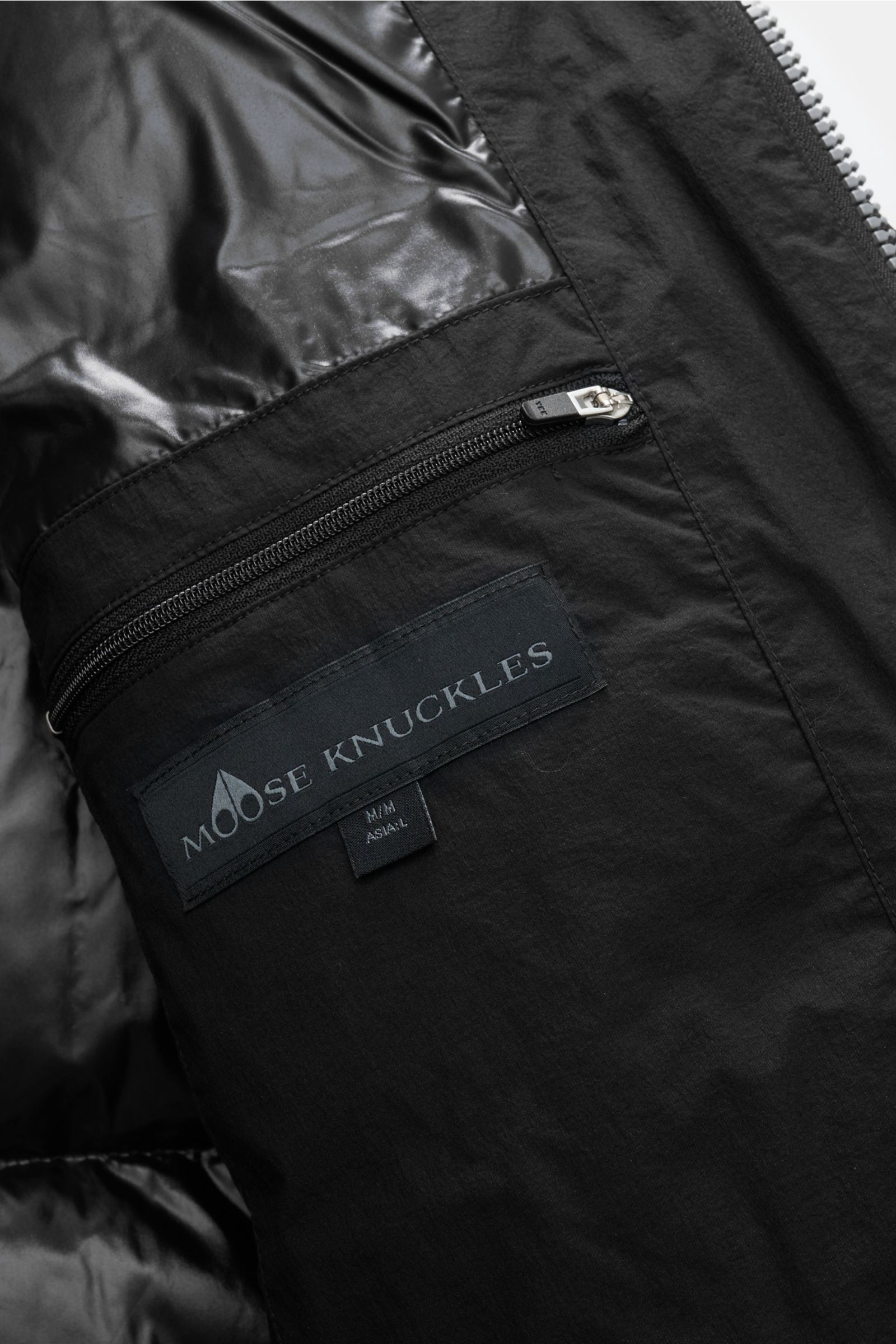 Nahaufnahme von der innenliegenden Reißverschlusstasche mit Moose Knuckles Label des schwarzen Moose Knuckles Daunenmantel 'Everest Long Puffer', wind- und wasserabweisend mit glänzendem Futterstoff.
