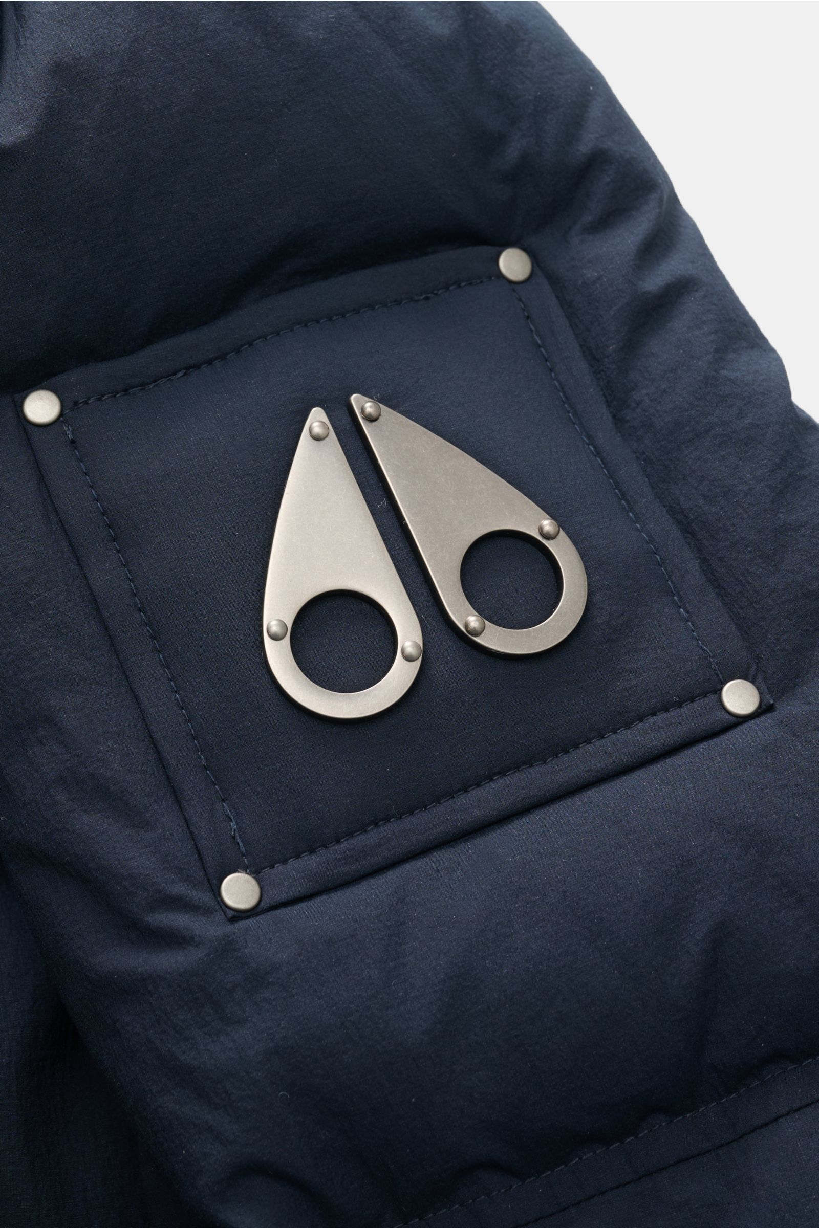 Nahaufnahme des typischen Logos am Oberarm des Moose Knuckles Daunenmantels 'Everest Long Puffer' navy aus hochwertiger, wind- und wasserabweisender Microfaser.