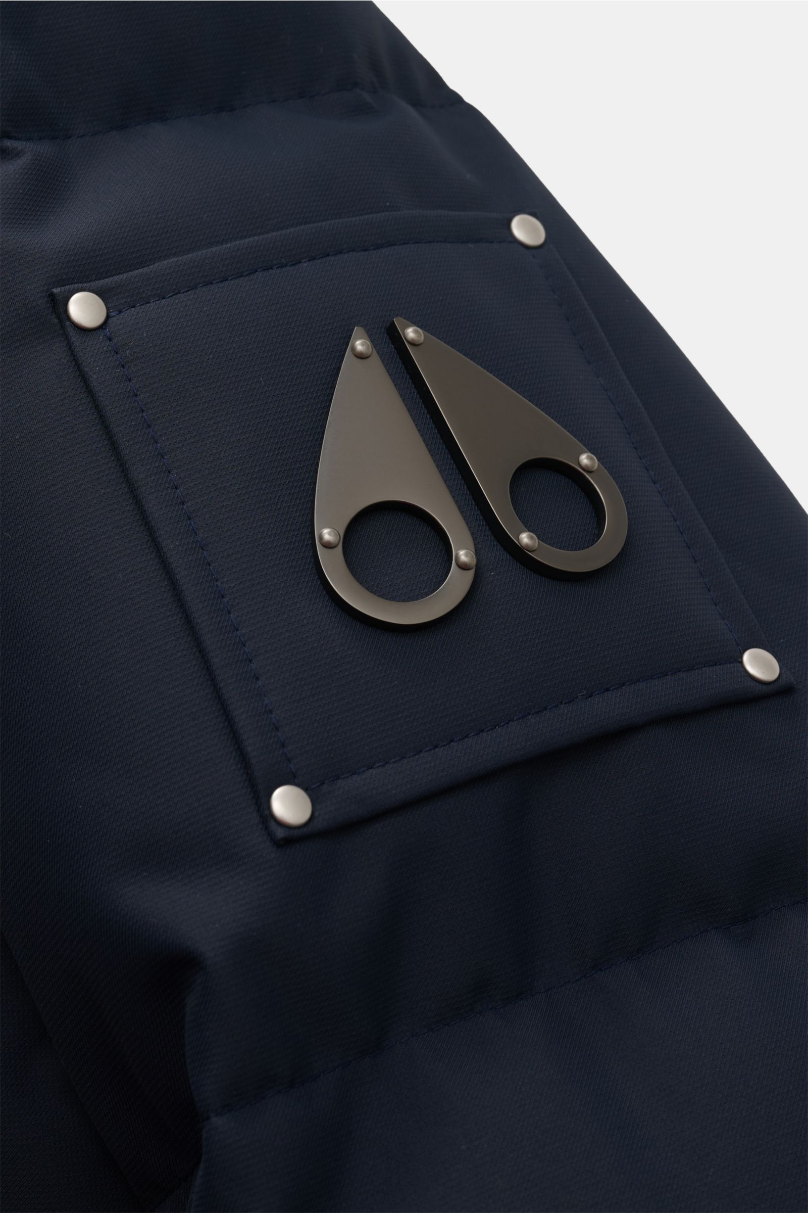 Nahaufnahme des Metall-Logos auf dem Oberarm der Moose Knuckles Daunenjacke 'Cloud 3Q' navy aus hochwertiger, wind- und wasserabweisender Microfaser.