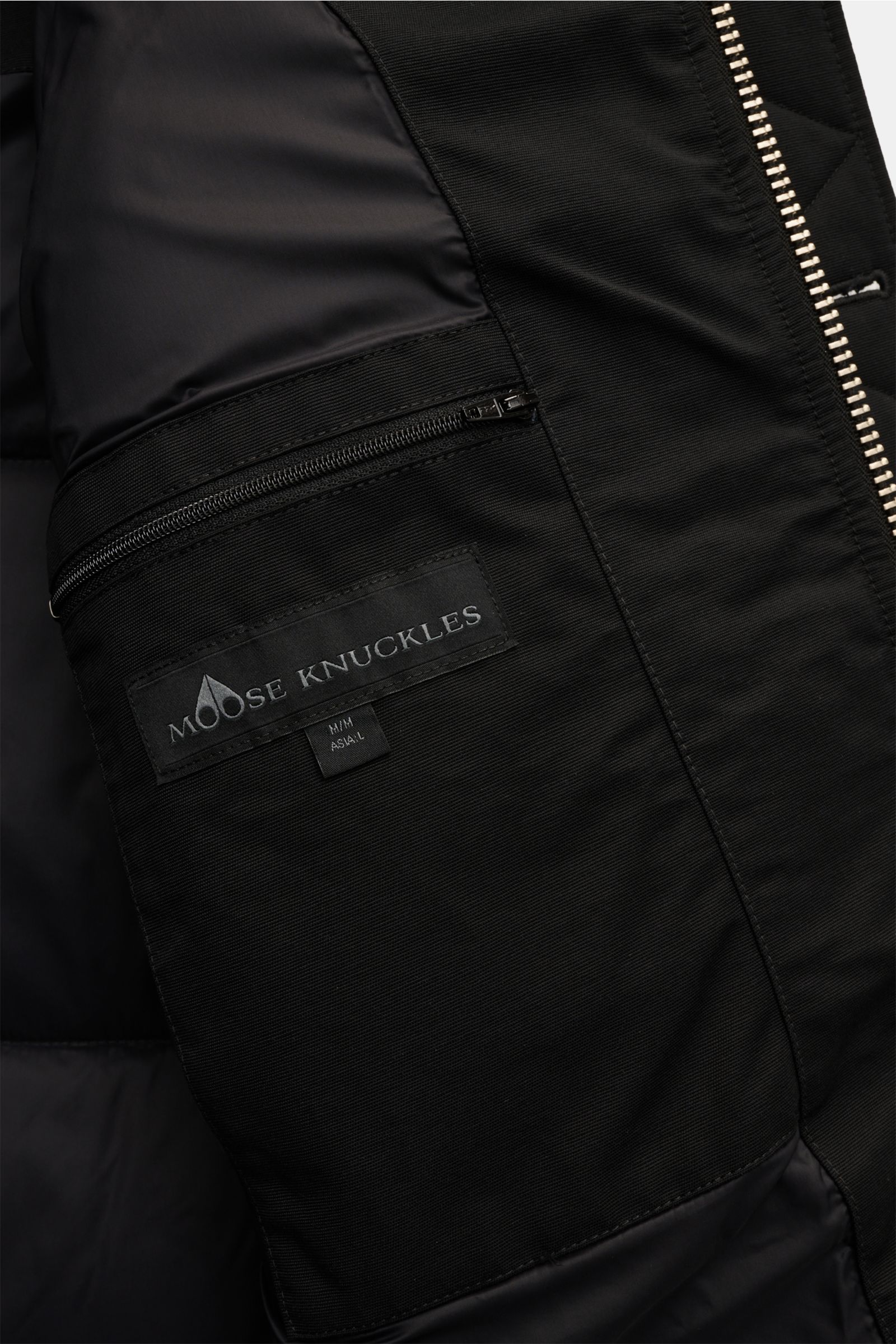 Close-up interior view of the black Moose Knuckles Daunenjacke 'Original 3q Neoshear' showing the zipped inner pocket with the Moose Knuckles logo label. Trotzt mühelos selbst extremer Kälte: Die MOOSE KNUCKLES Jacke sorgt mit ihrer hochwertigen 90/10-Dau