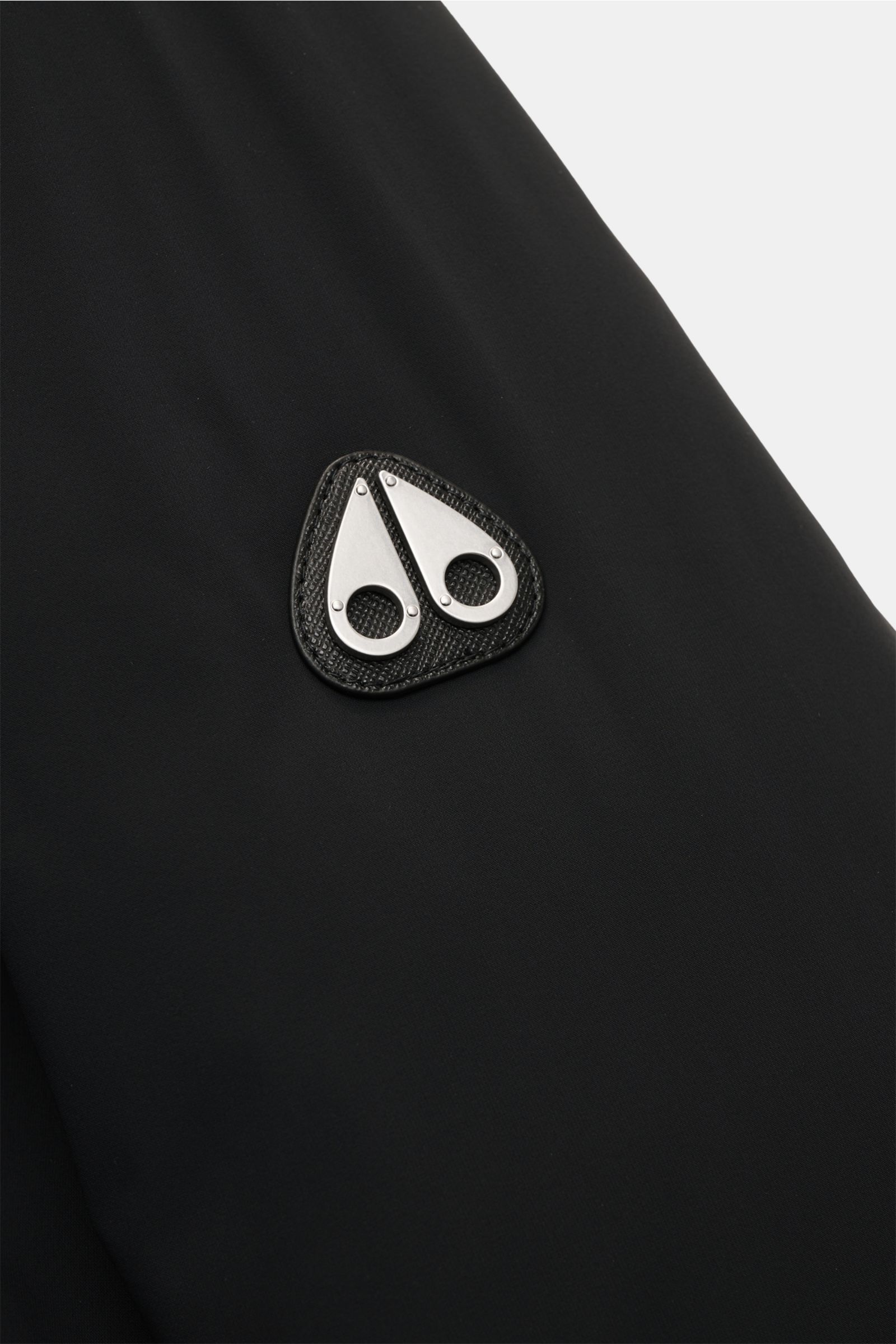Nahaufnahme des Metall-Logos am Oberarm der schwarzen Moose Knuckles Daunenjacke 'Northdale Hoodie' aus glatter, wind- und wasserabweisender Microfaser.