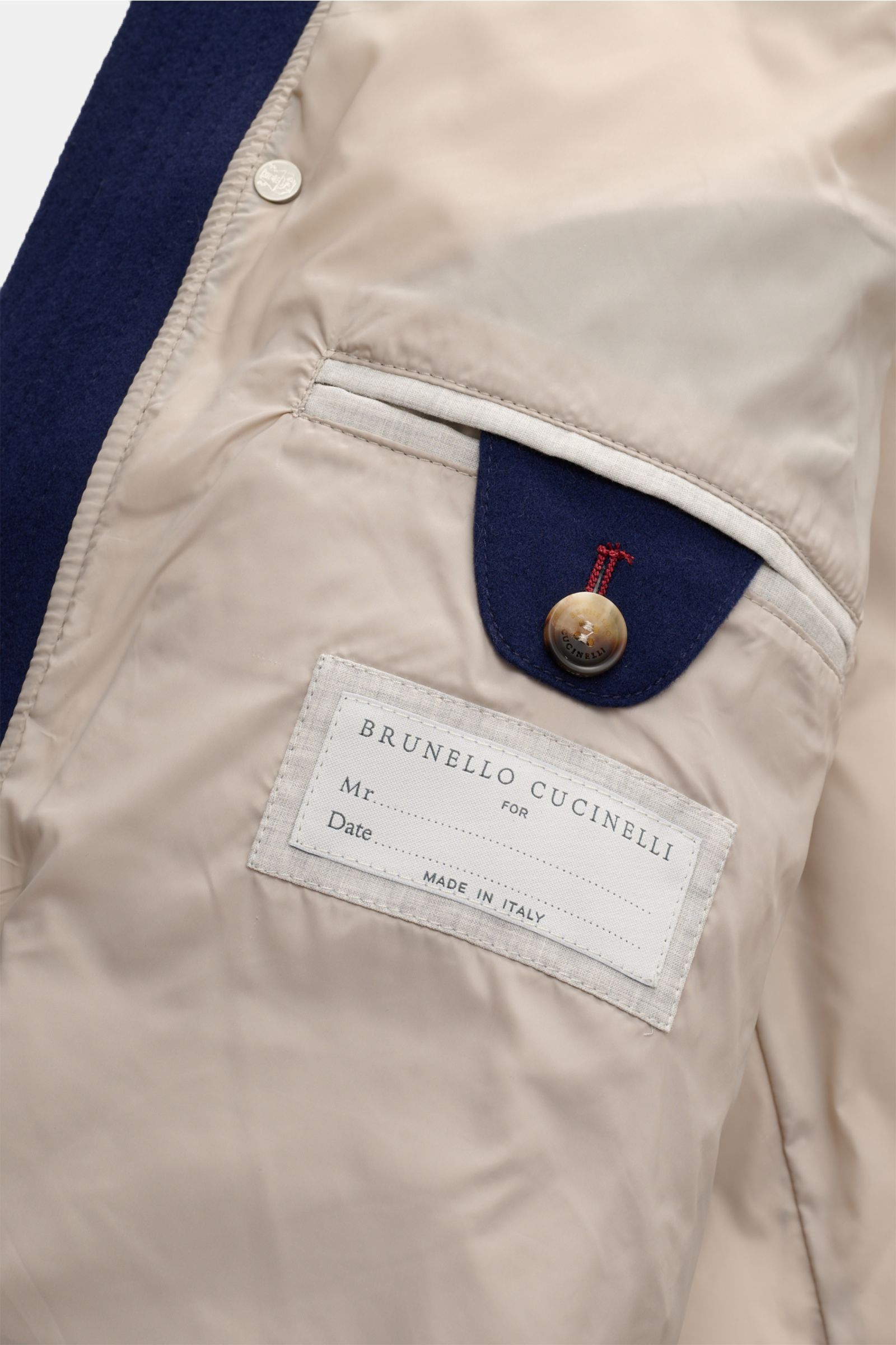Alt-Tag (124 Zeichen): Nahaufnahme der Innenseite der Brunello Cucinelli Daunenjacke dunkelblau mit Label, Knopf und beige Futterstoff.