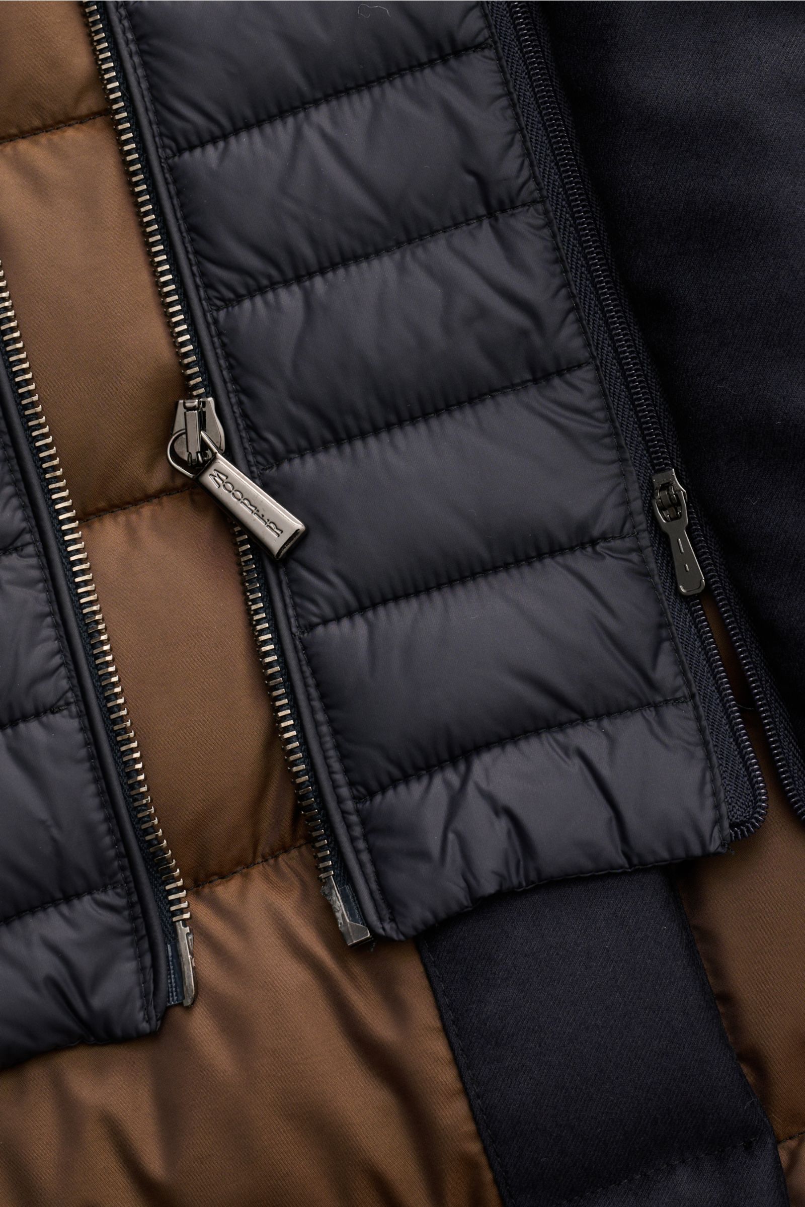 Close-up image of the navy Moorer Daunenjacke 'Calegari' showing its water-repellent outer material, quilted design, and metal zippers with the brand name, photographed from above. Stilvoll jeder Wetterlage trotzen wir mit der langen Jacke 'Calegari' aus 