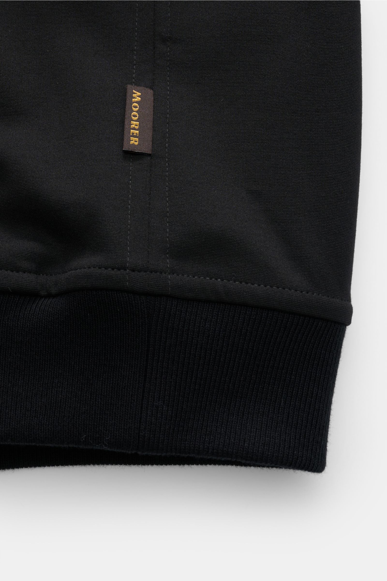 Close-up front view of the ribbed cuff and fabric tag of the Moorer Blouson 'Astaire' schwarz, a slim fit jersey-blouson made of viscose and microfiber, featuring soft touch, two-way zipper, rib-knit collar, piped front pockets, comfortable rib cuffs, and