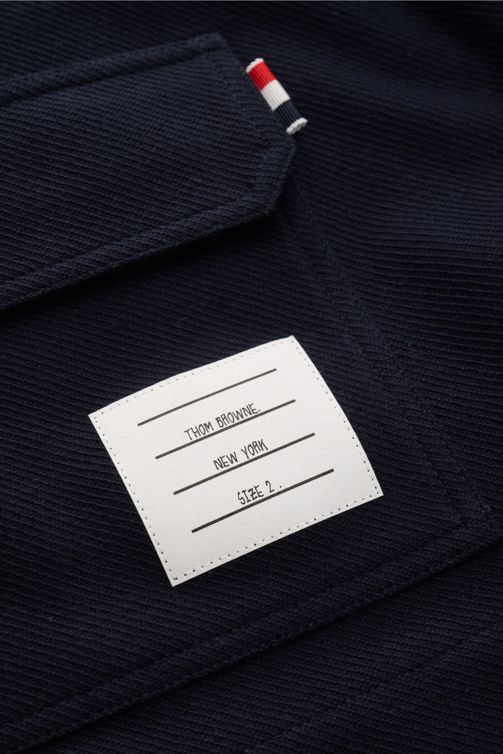 Close-up of Thom Browne Jacke navy showing textured cotton fabric, label with brand and size, and signature tricolor tab on pocket edge, photographed from above.

Description for reference: 
Legeres Layering-Piece: Die THOM BROWNE Jacke besticht mit sport