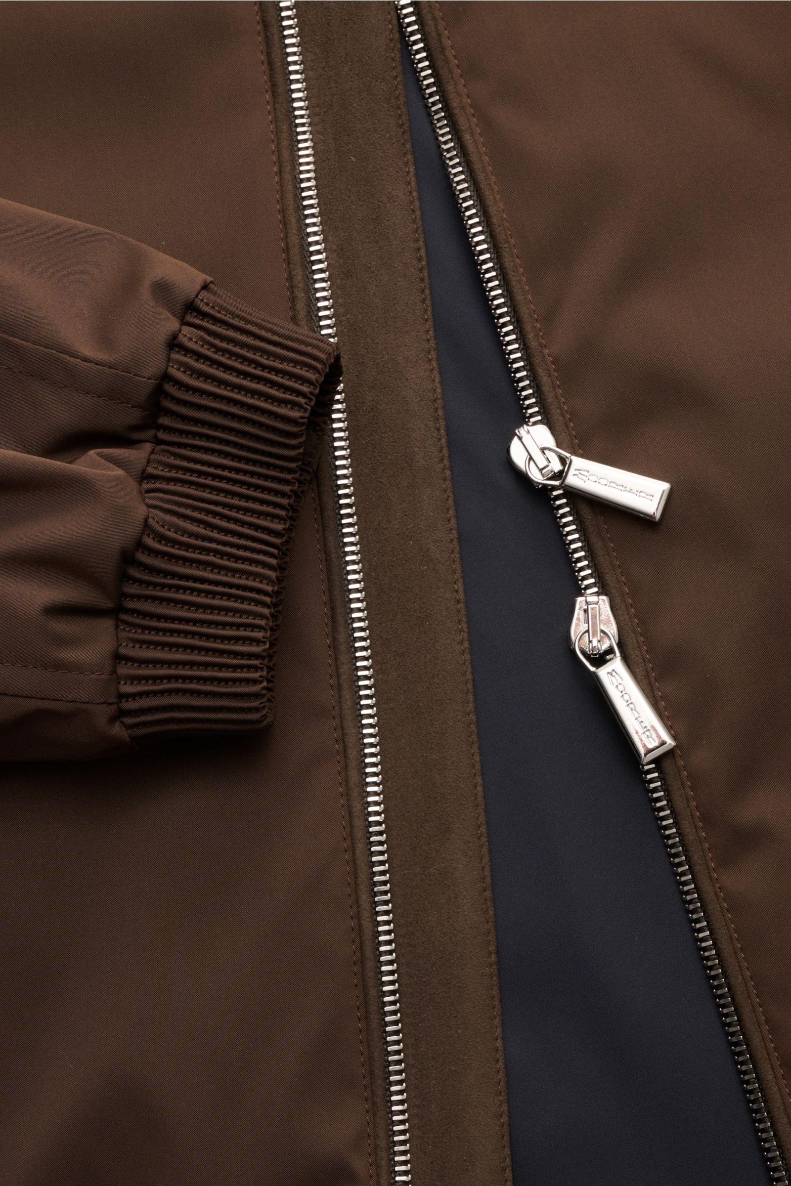 Close-up front view of the zipper and cuff of the Moorer Blouson 'Albori TJ' dunkelbraun, featuring high-quality microfiber, smooth grip, water-repellent, slim fit, hood with drawstring, zipper pockets, elastic hem and cuffs, smooth lining, and two inner 