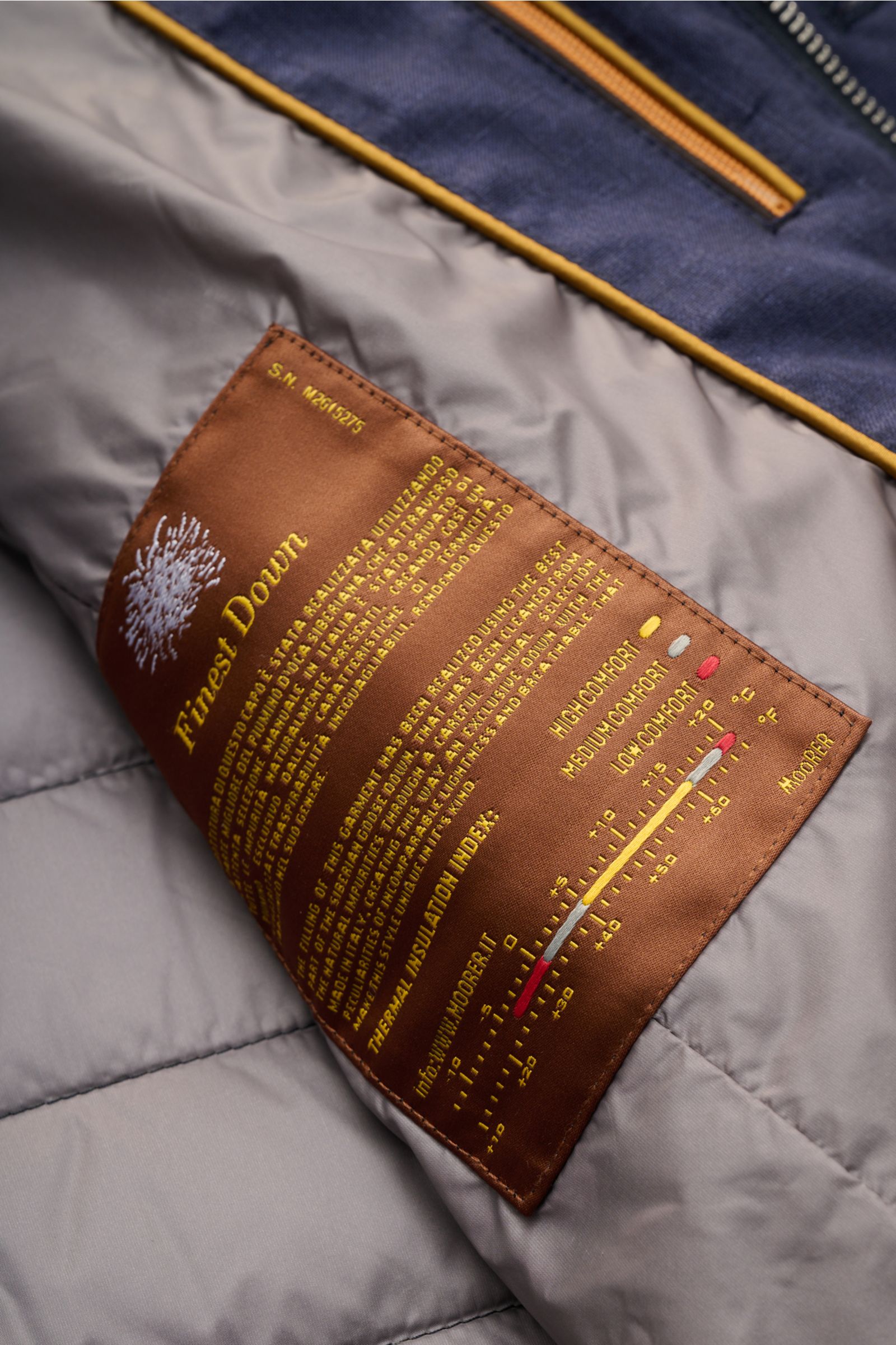 Close-up interior view of the Moorer Down jacket 'Zavyer' navy mottled showing a brown label with yellow text detailing finest down insulation and thermal index, smooth lining, and quilted stitching. The elegant quilted jacket 'Zavyer' by MOORER will see 