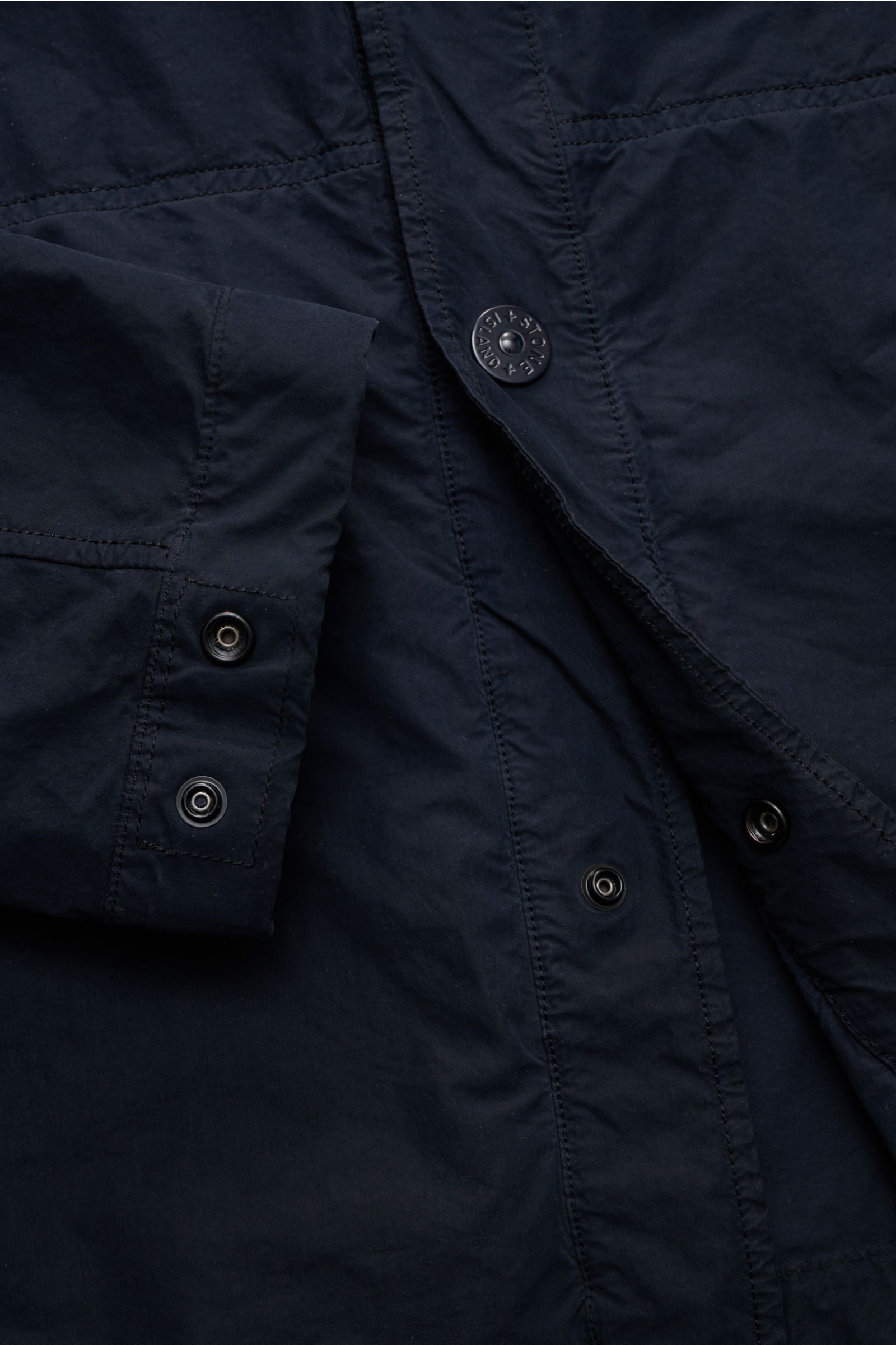 Close-up front and sleeve view of Stone Island Jacke 'Ghost Piece' navy, showing garment dyed microfiber, snap button placket, cuff buttons, and tonal compass badge. Urbanes Essential: Die STONE ISLAND Jacke aus der 'Ghost Collection' überzeugt mit legere