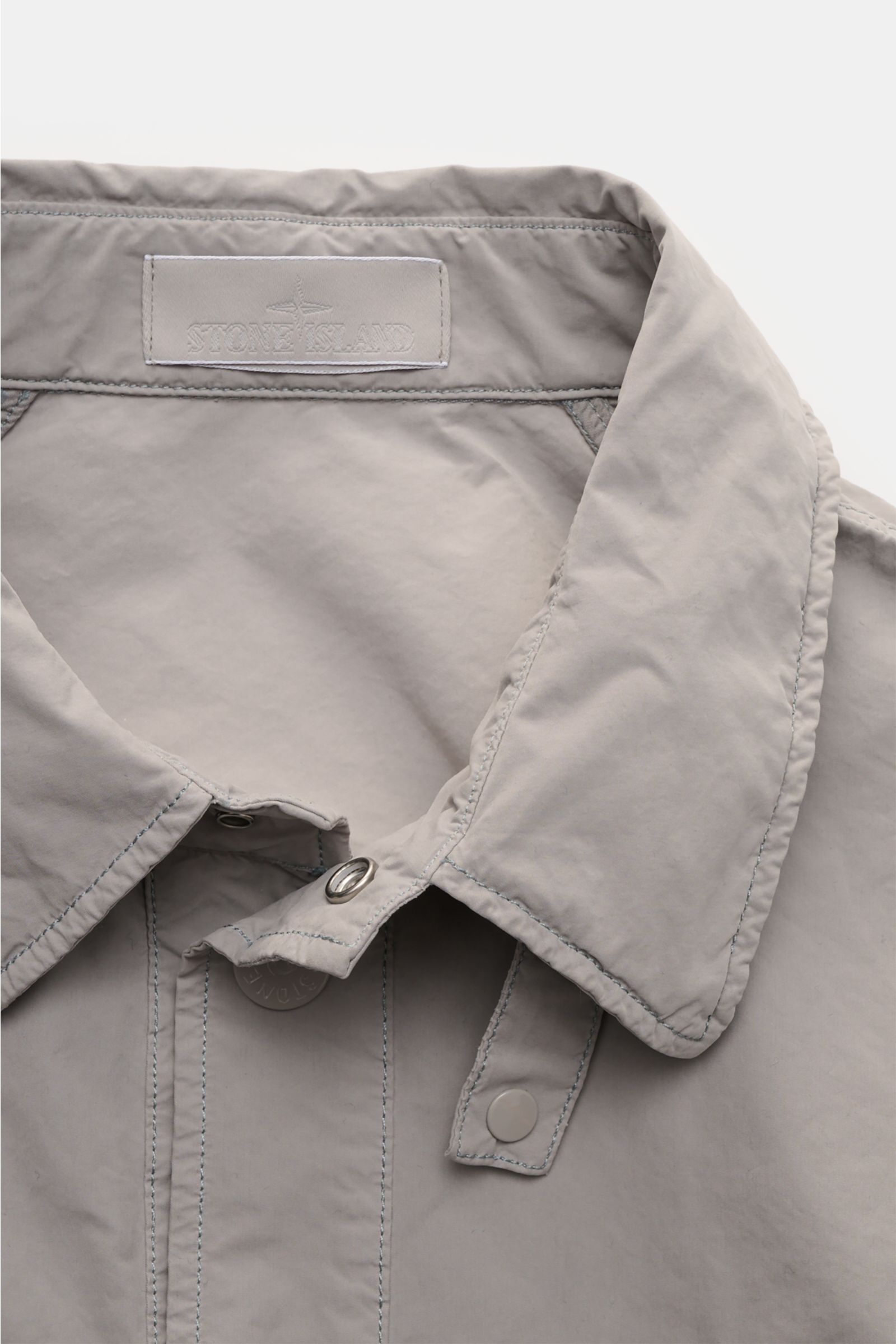 Stone Island Jacket 'Ghost Piece' light grey shown close-up from above, featuring high-quality microfibre, soft feel, snap button placket, and turn-down collar with buttoned tab. Urban essential from the 'Ghost Collection' with slim fit, front pockets wit