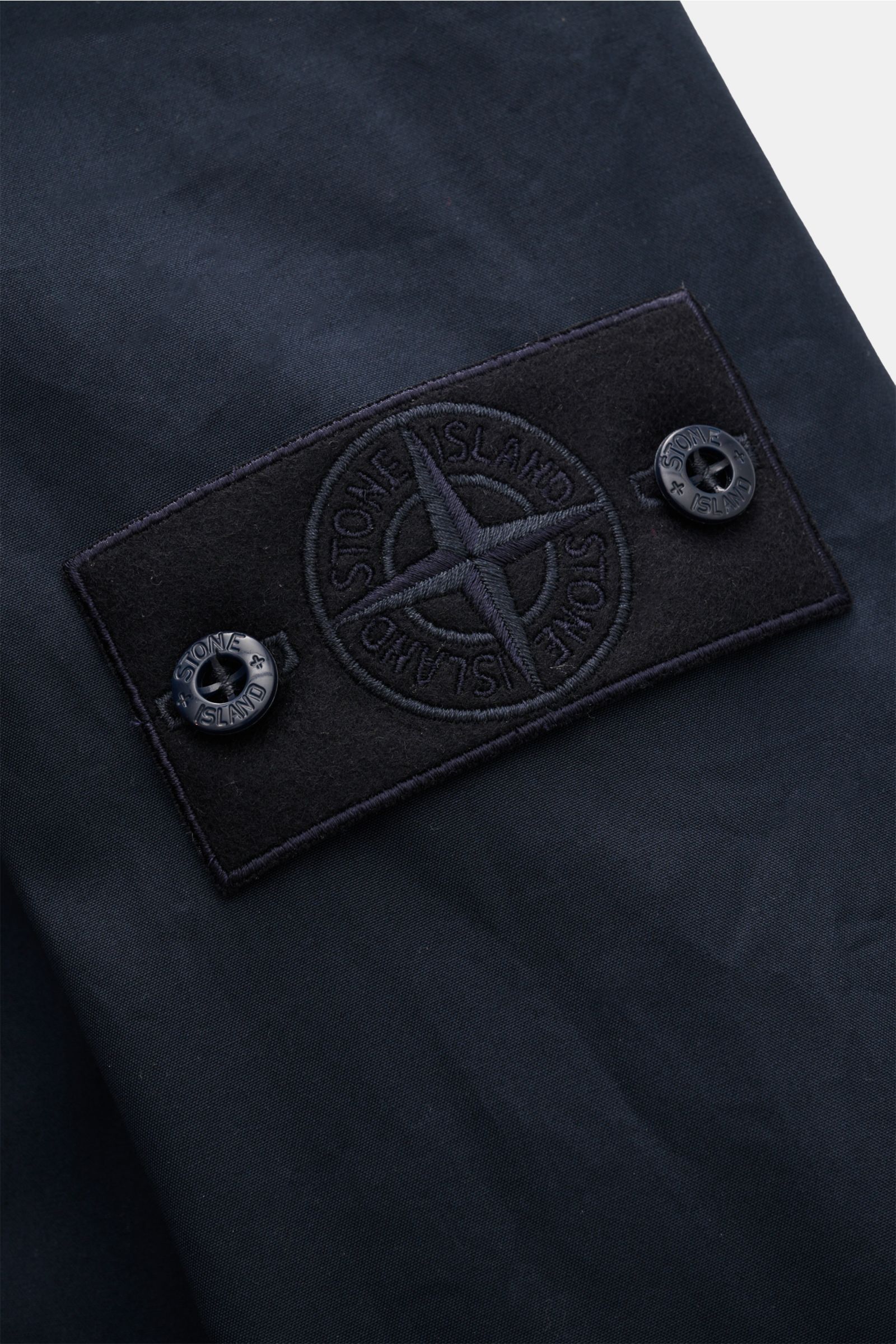 Close-up side view of the Stone Island Jacke 'All-Weather S.I. Ghost Piece' navy showing the black embroidered logo patch on navy cotton canvas.

Description text to include:  
Funktion trifft Komfort: Die STONE ISLAND Jacke aus der 'Ghost' Capsule wurde 