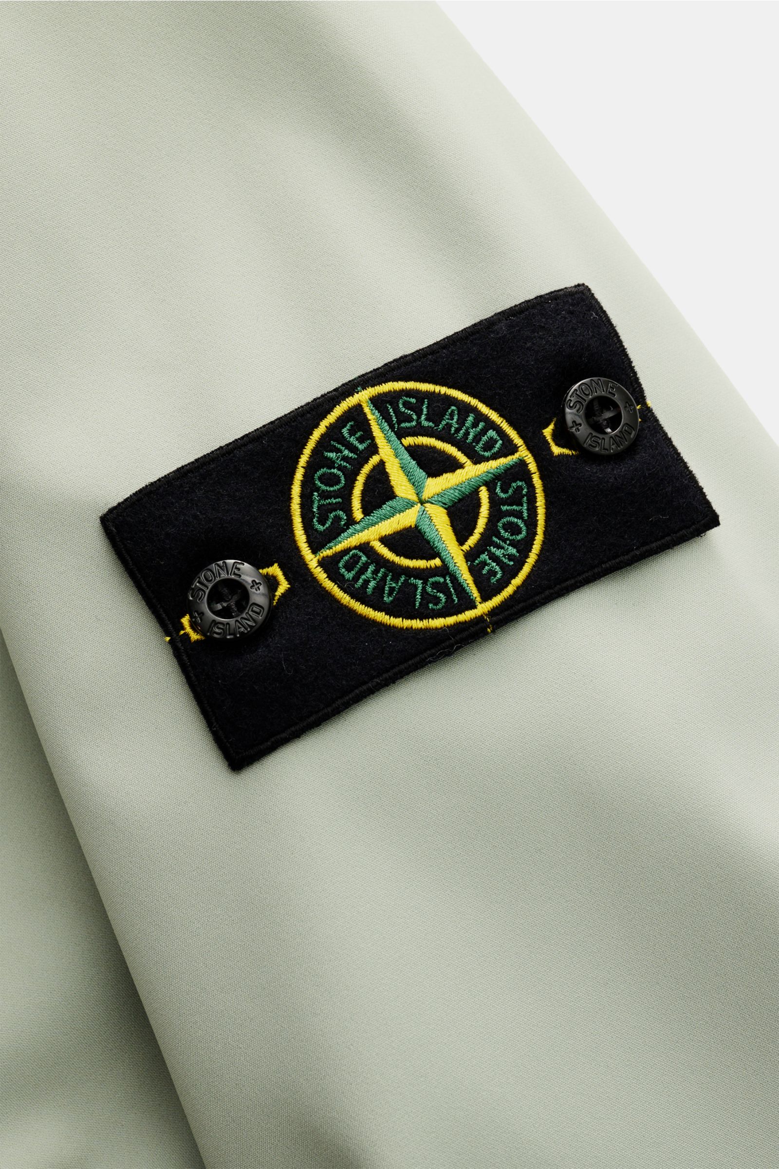 Nahaufnahme des Kompass-Badges am Oberarm des Stone Island Blouson 'Light Soft Shell-R' salbei aus hochwertiger, wind- und wasserabweisender Microfaser.