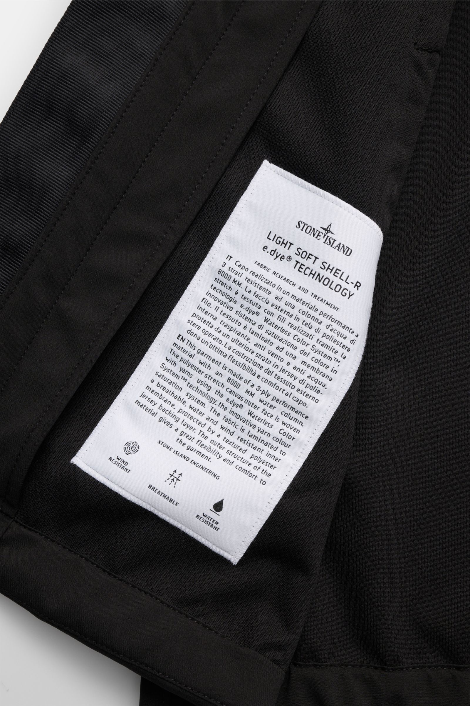 Nahaufnahme des Innenfutters der Stone Island Jacke 'Light Soft Shell-R' schwarz, zeigt weißes Etikett mit Material- und Technologieinfos, geschmeidiger Futterstoff, wind- und wasserabweisend, Slim Fit, elastische Bündchen, Kompass-Badge, Fronttaschen mit