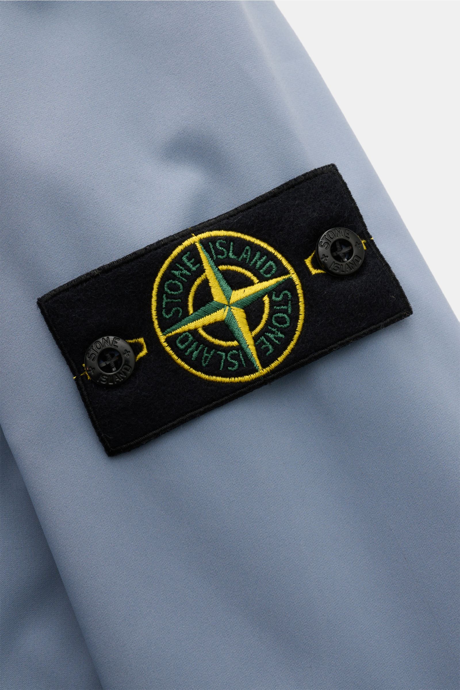 Nahaufnahme der Kompass-Badge an der Ärmelmanschette der Stone Island Jacke 'Light Soft Shell-R' rauchblau aus hochwertiger, wind- und wasserabweisender Microfaser.