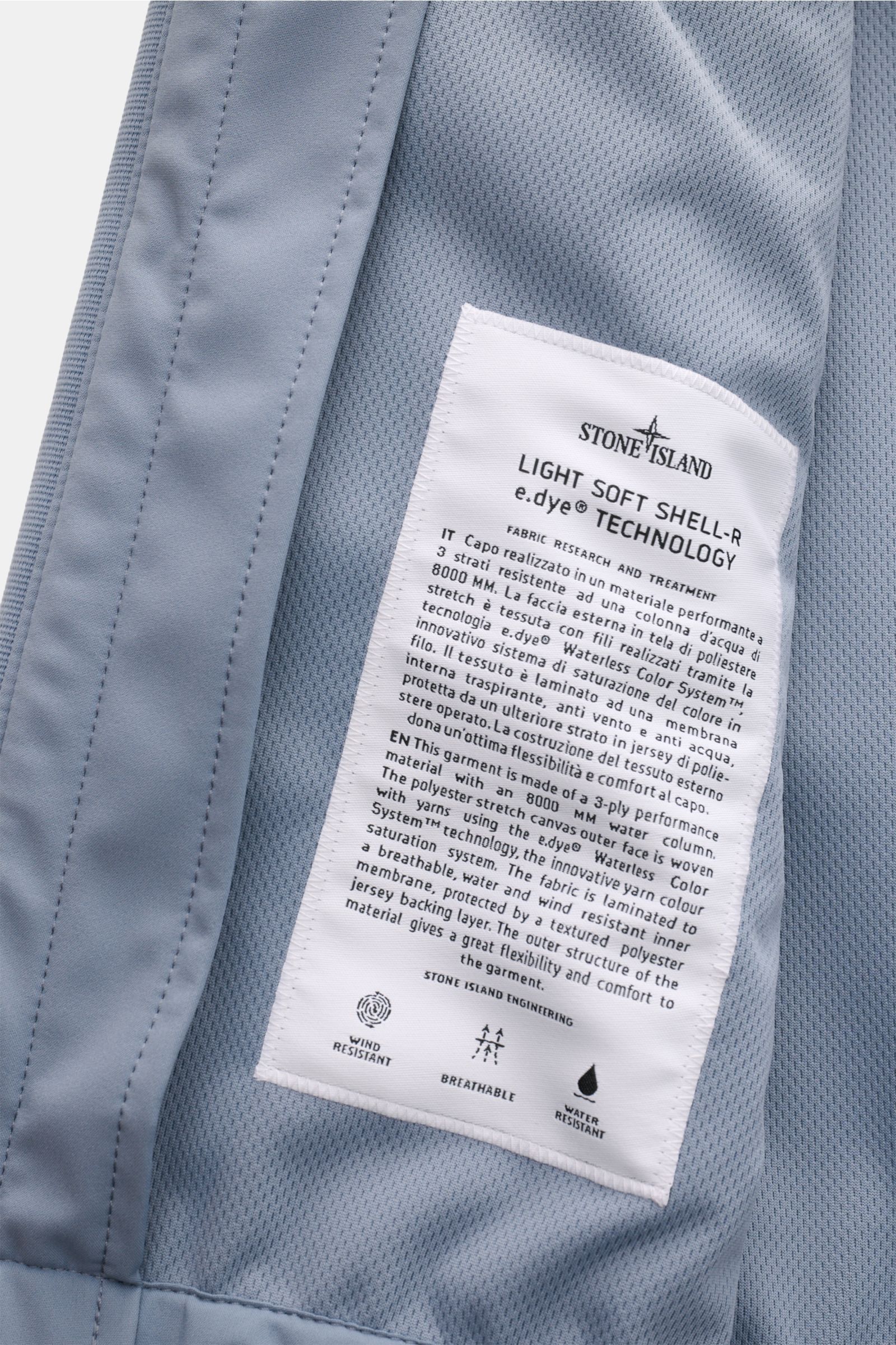 Nahaufnahme des Innenfutters der Stone Island Jacke 'Light Soft Shell-R' rauchblau mit weißem Label, das Material und Funktion beschreibt.