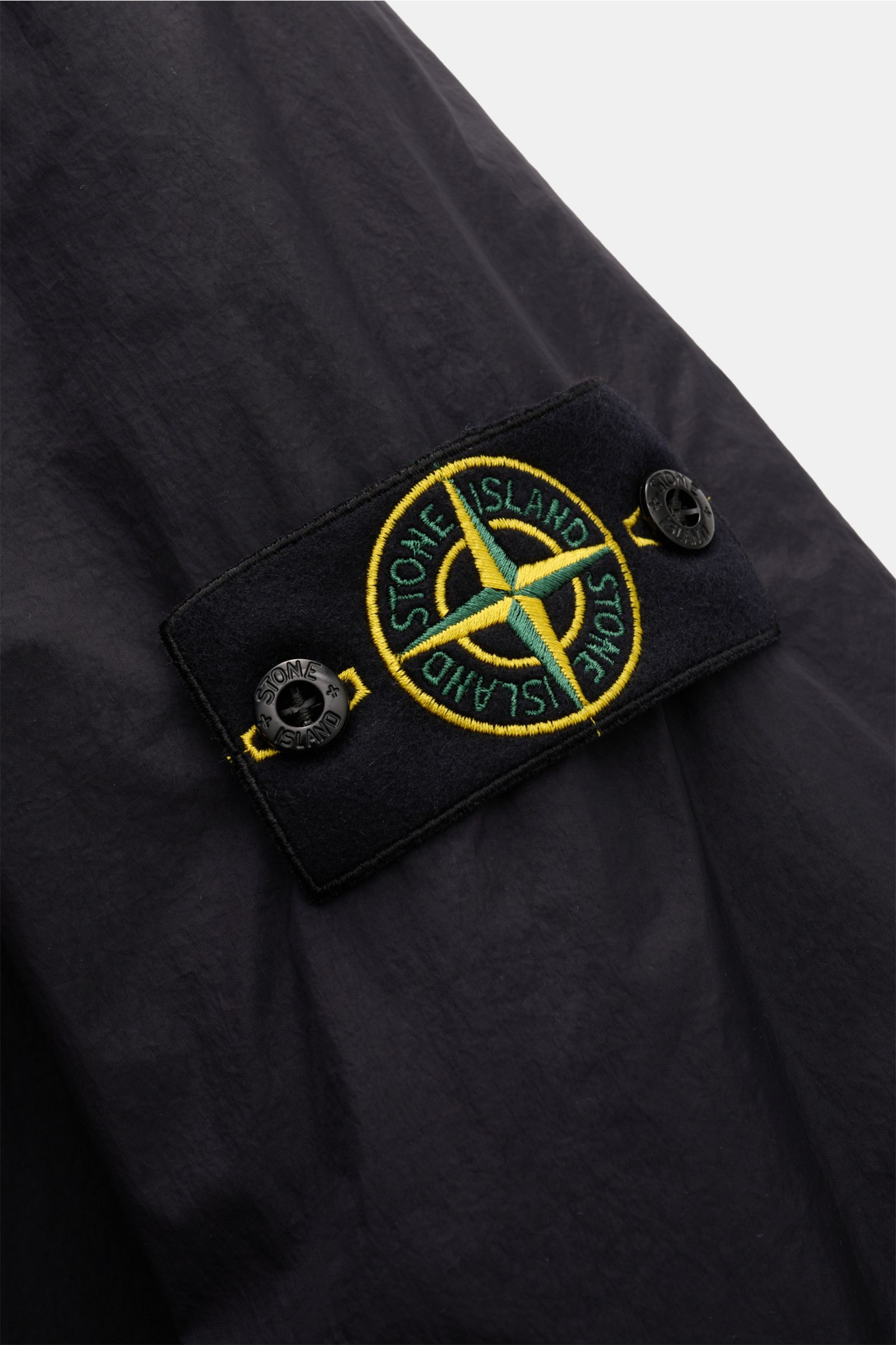 Close-up from a side angle of the Stone Island Jacke 'Garment Dyed Crinkle Reps NY' dark navy, showing the embroidered compass badge on the sleeve with black buttons on lightweight, crinkled microfiber fabric.

The jacket features:
- high-quality microfib