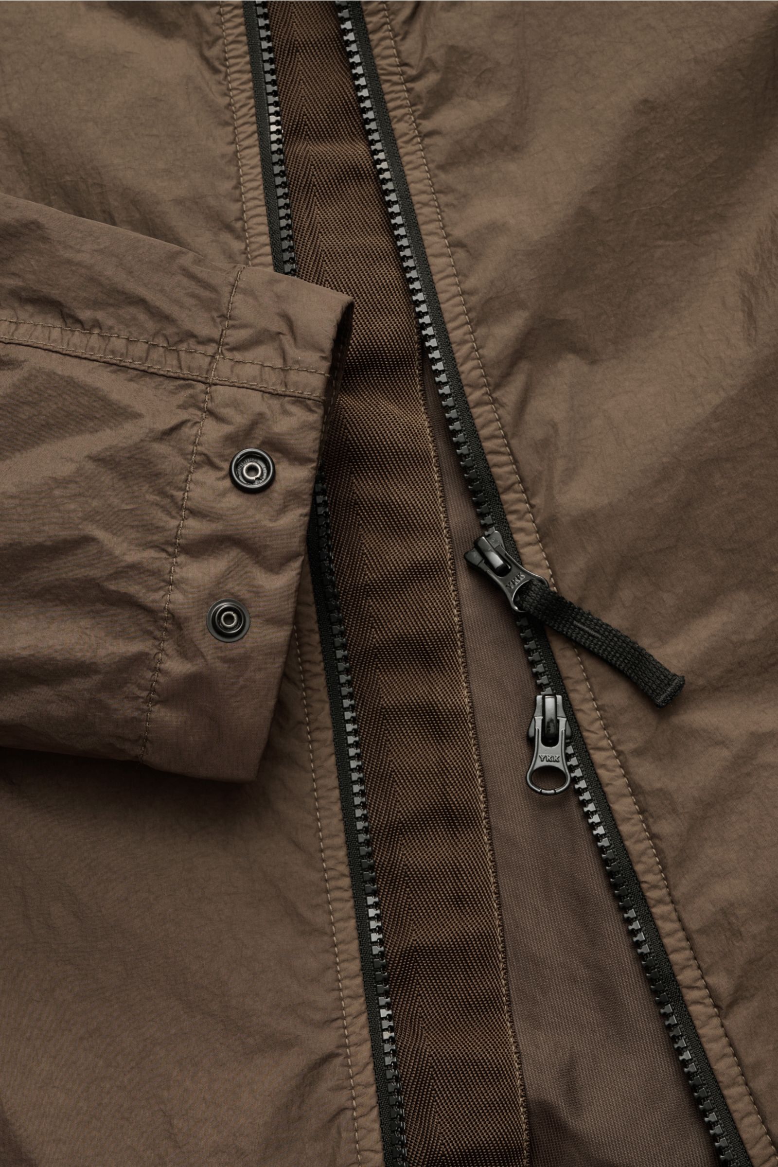 Nahaufnahme der wasserabweisenden Stone Island Jacke 'Garment Dyed Crincle Reps NY' braun mit Reißverschluss und Druckknopf am Ärmel.